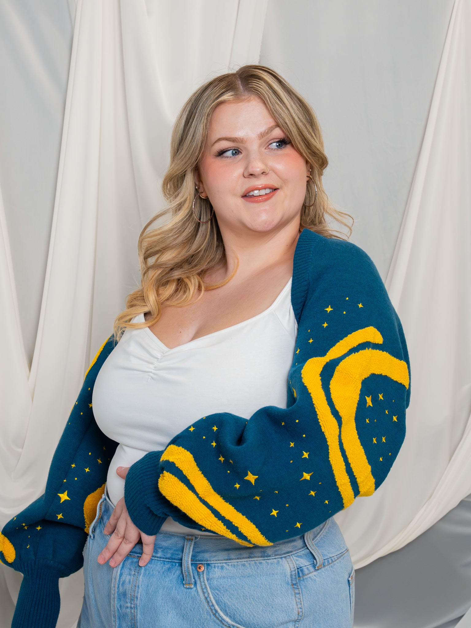 Woman wearing a blue and yellow cardigan with a white top and jeans against a white curtain background