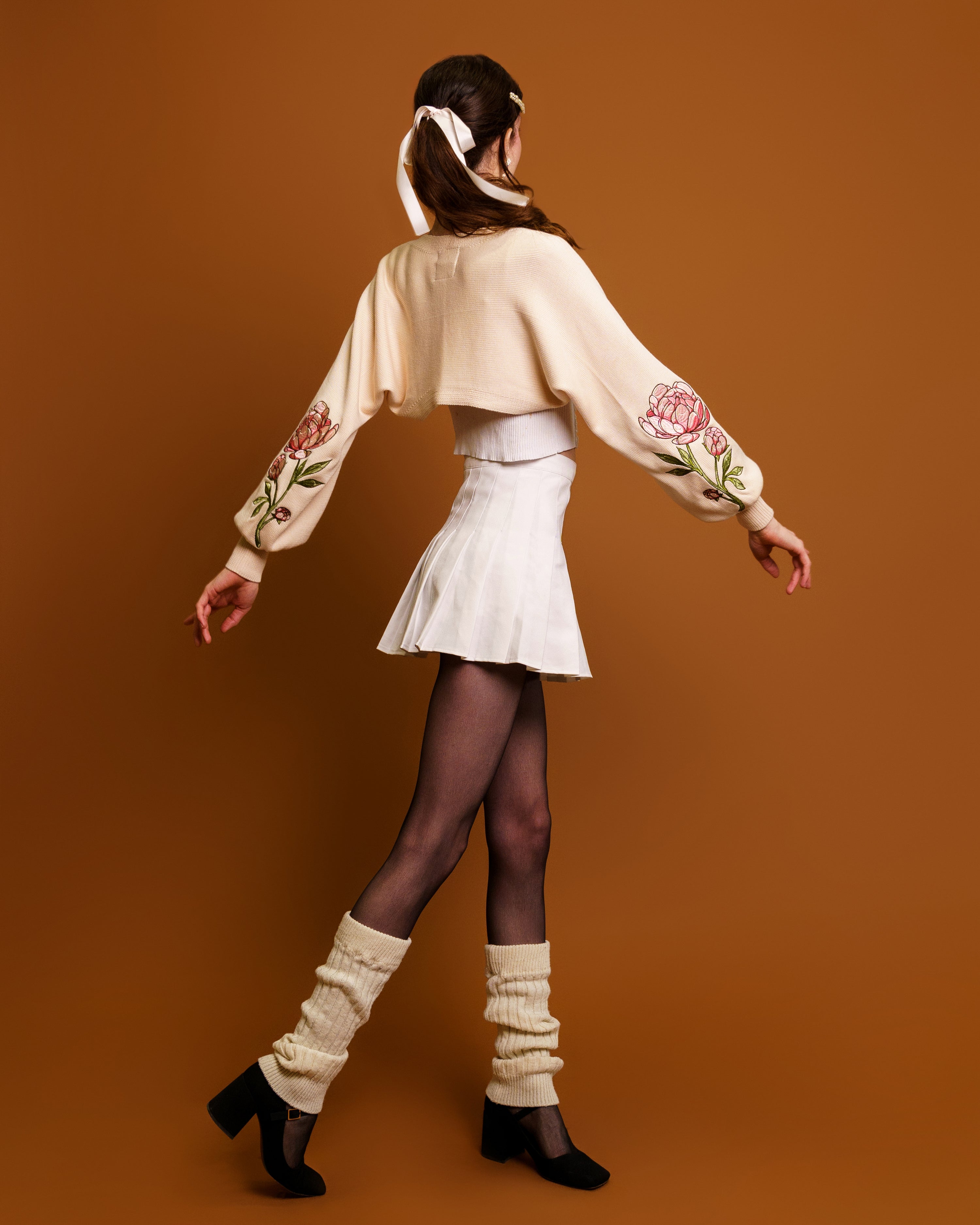 Woman wearing bashful cream bolero in size extra small swaying in front of orange background.