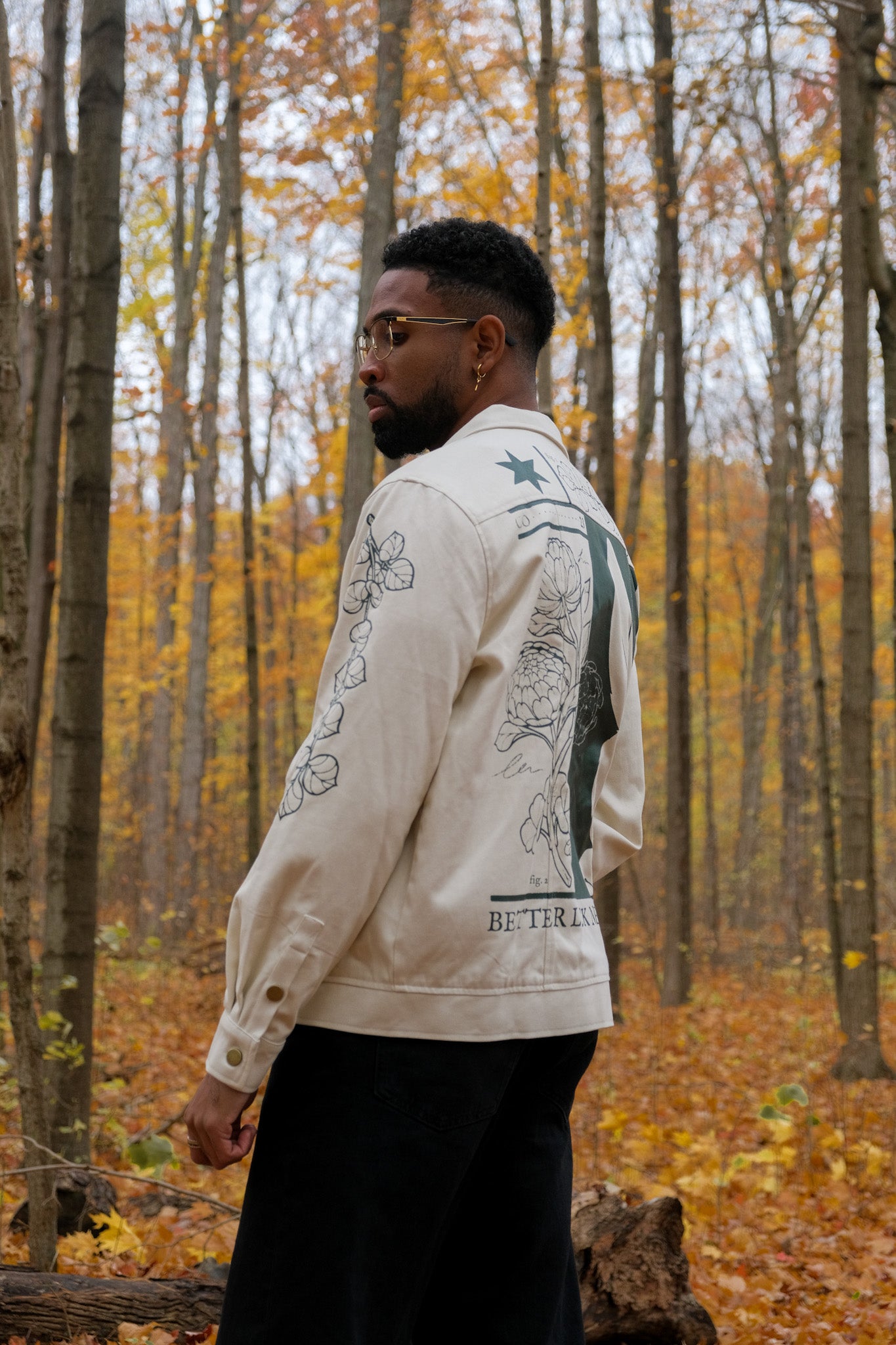 Man wearing white better luck next time jacket in size large standing slightly to the side, showing green leaf graphic on sleeve in a forest.