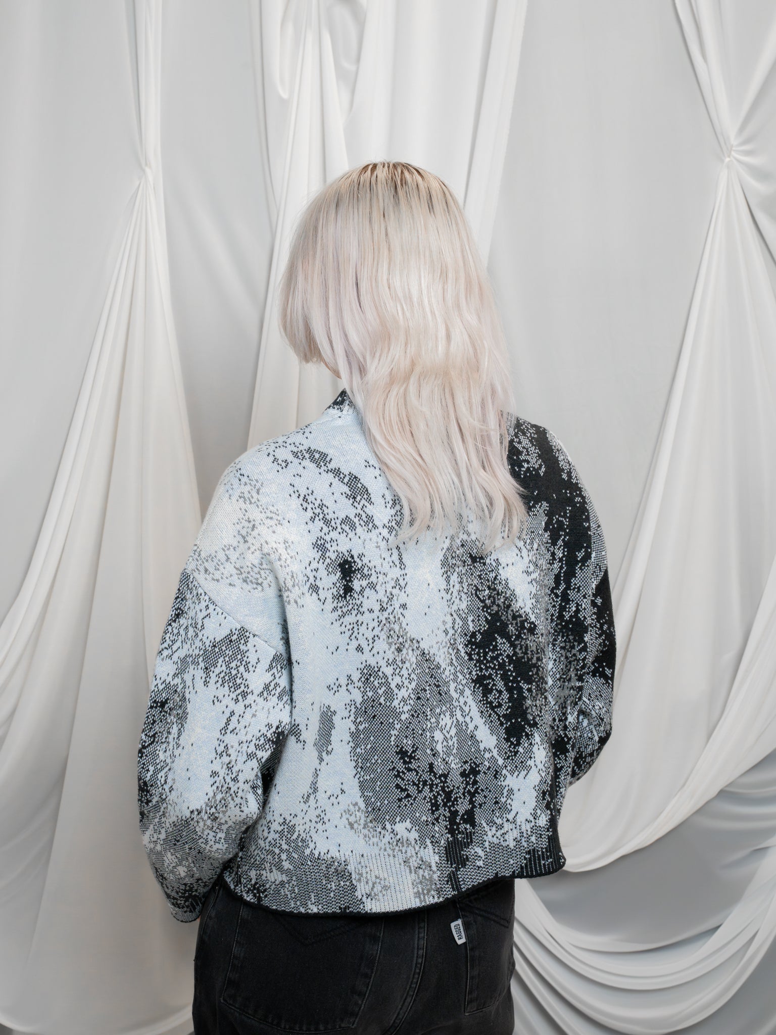 Back view of woman wearing white, grey and black cropped knit lunar sweater in size extra small in front of white draped fabric. 