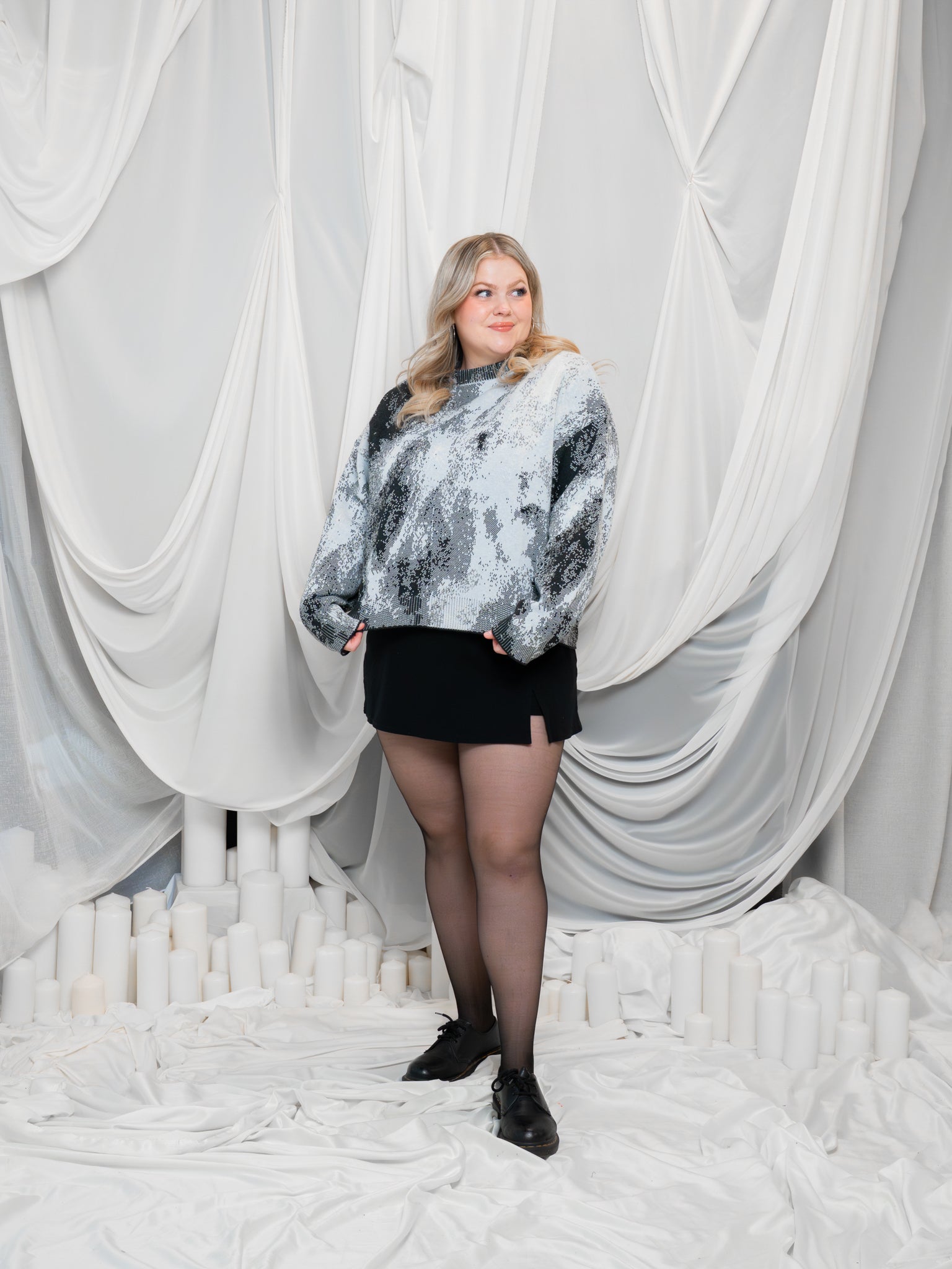 Woman wearing white, grey and black cropped knit lunar sweater in size large with black skirt in front of white draped fabric.