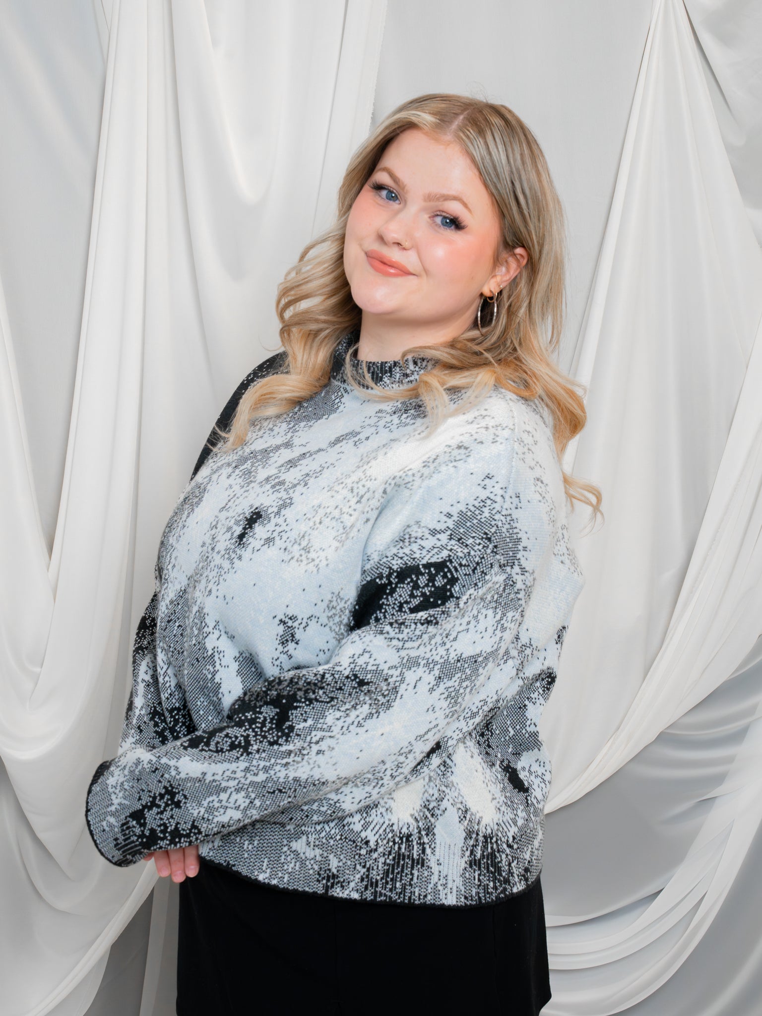 Woman wearing white, grey and black cropped knit lunar sweater in size large posing in front of white draped fabric.
