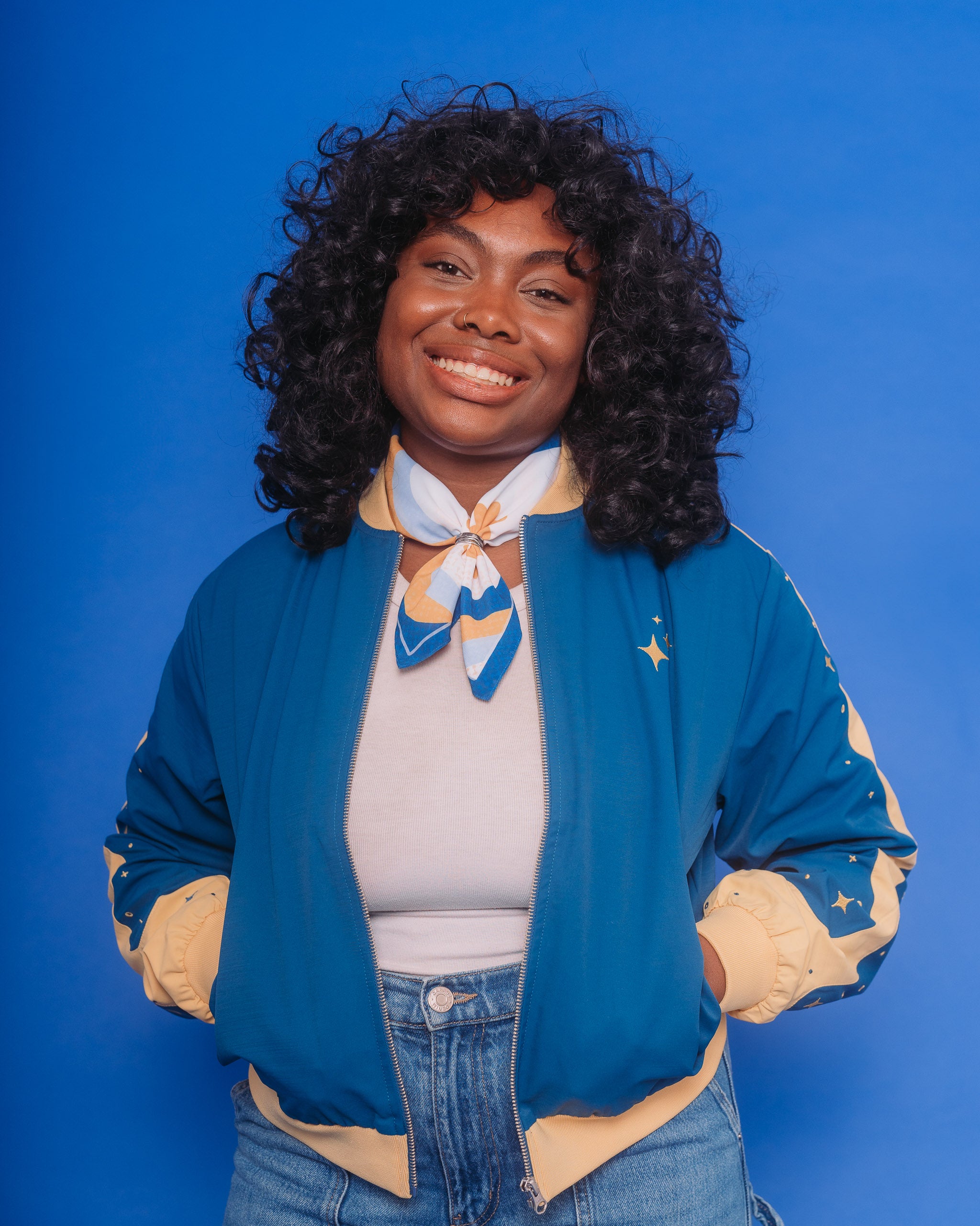 Woman wearing blue moon bomber jacket in size medium with hands in pockets in front of blue background.