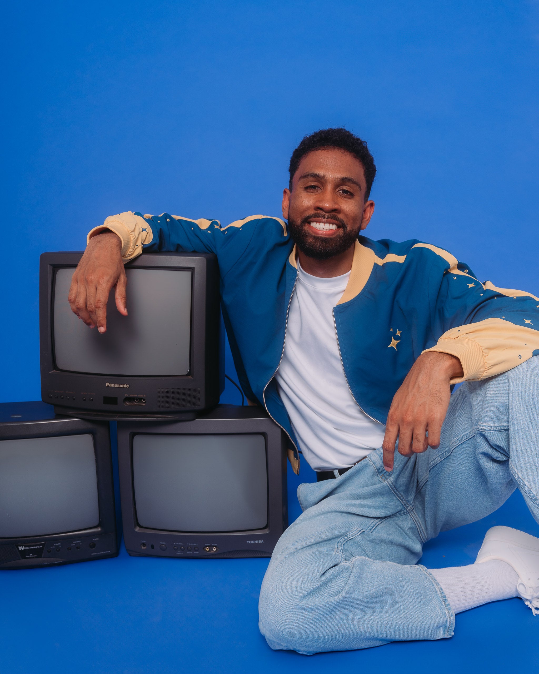 Man wearing blue moon bomber jacket in size extra large posing with vintage tvs on ground.