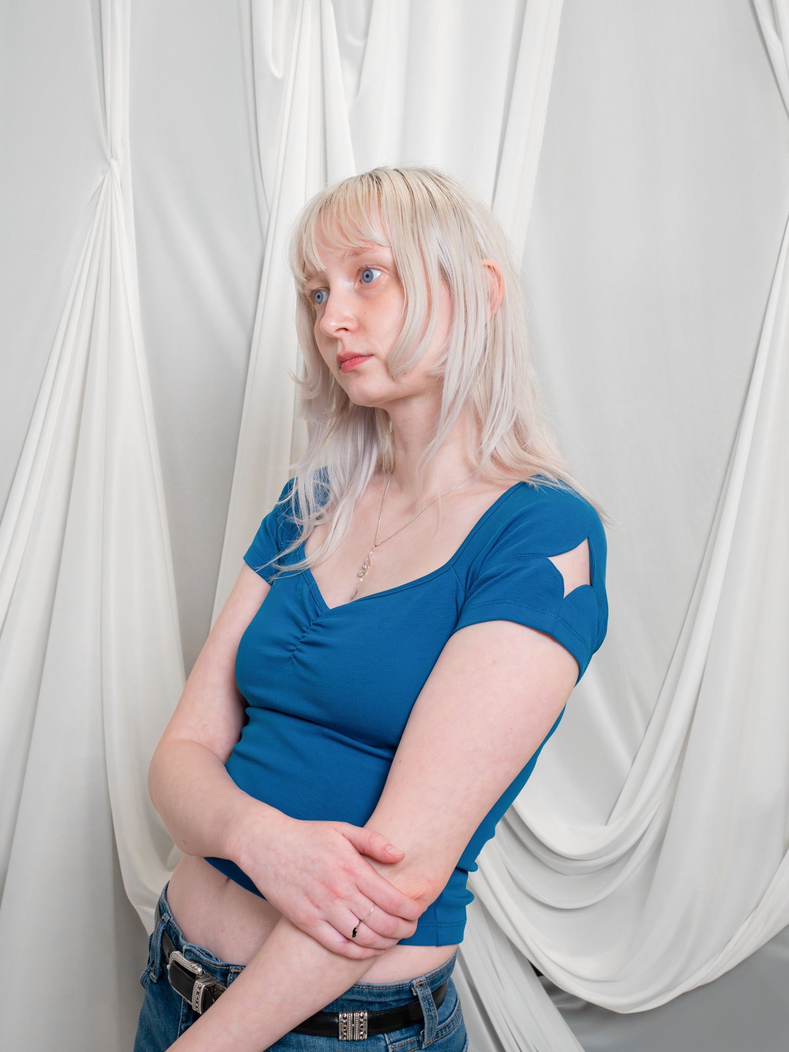 Three quarter view of woman wearing blue moon (blue) cap sleeve tee in size extra small with loves me not necklace in front of white fabric draped background.