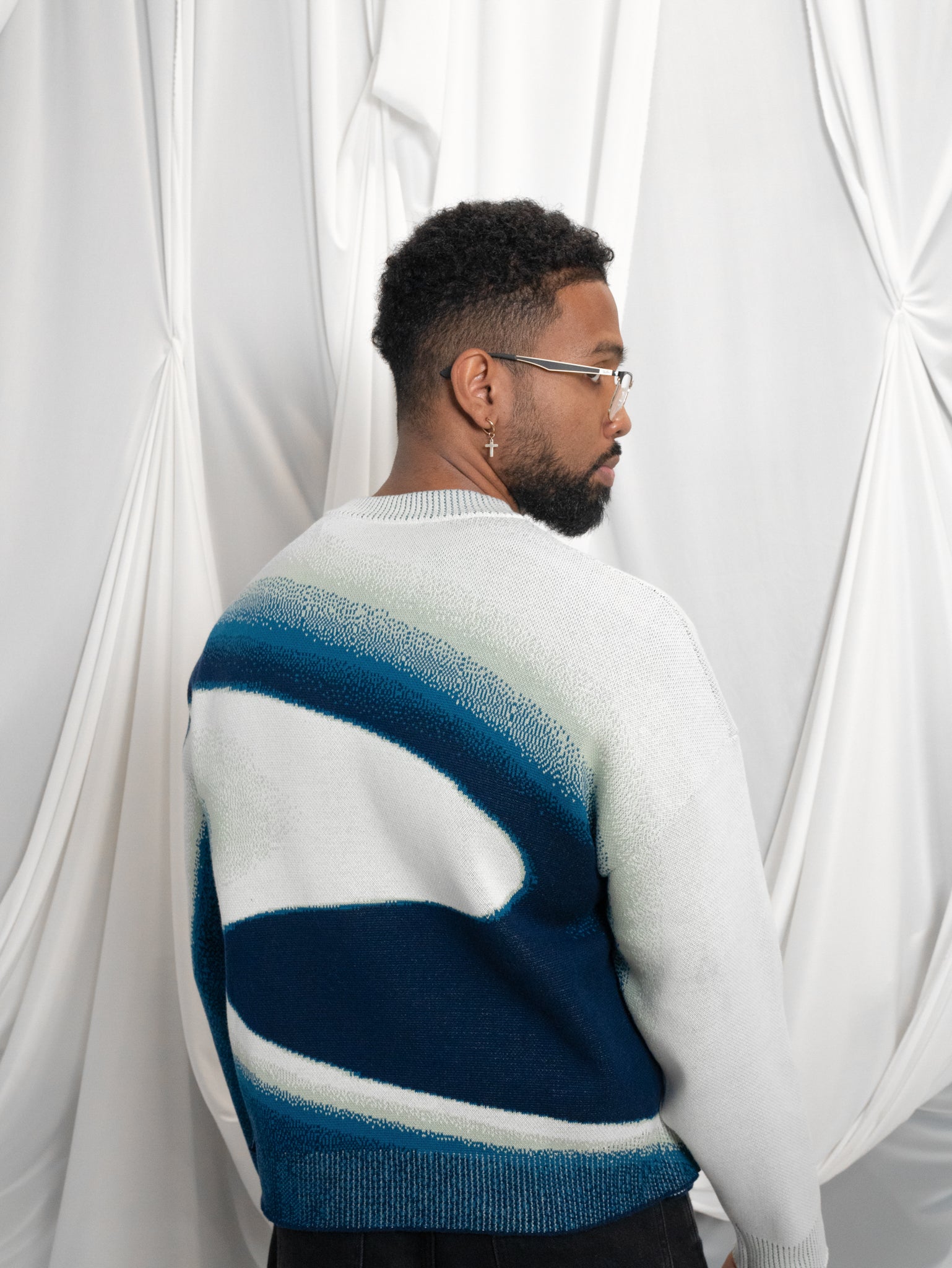 Back view of man wearing blue and white cropped knit stardust sweater in size large in front of white draped fabric.