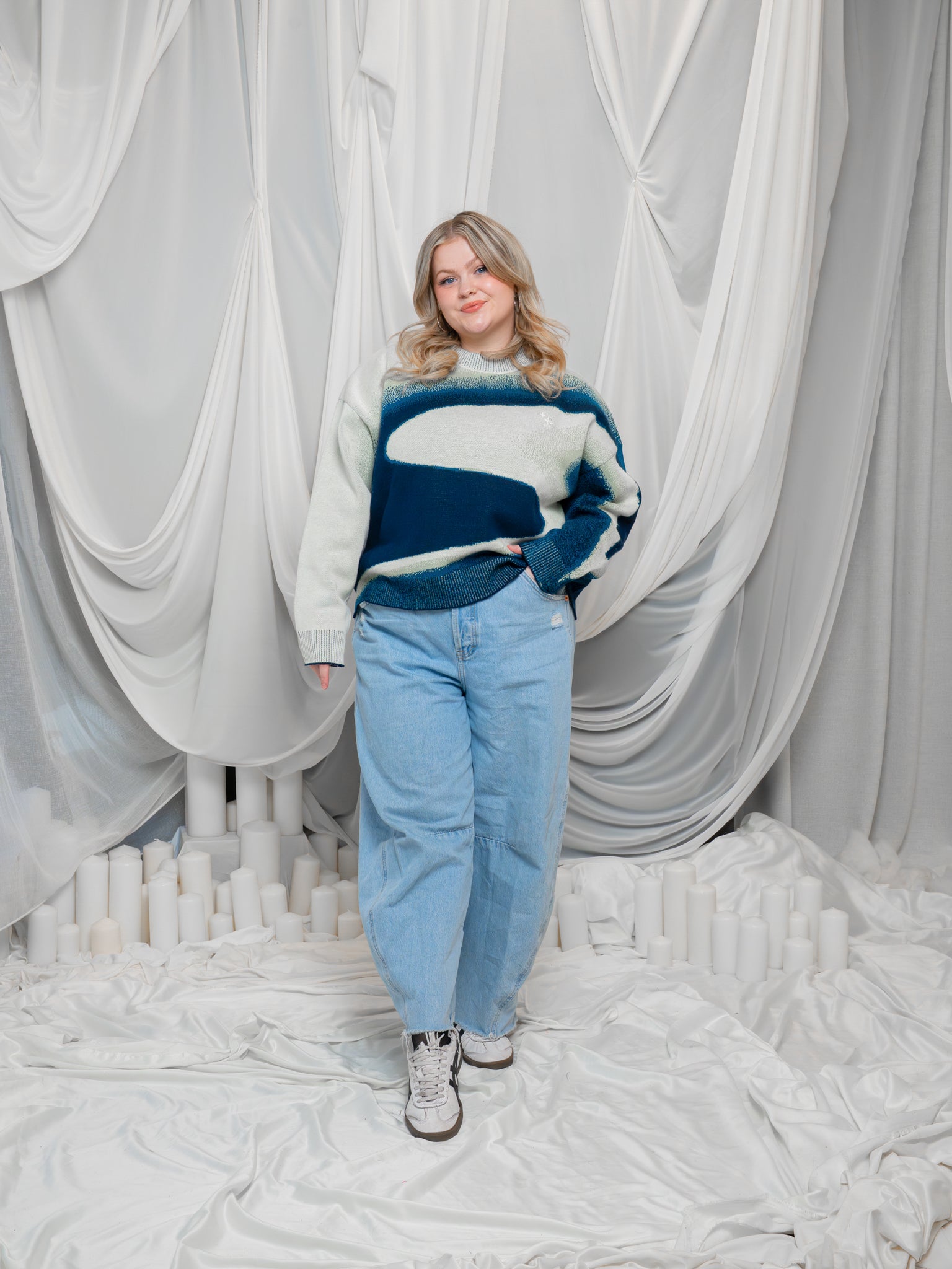 Woman wearing wearing blue and white cropped knit stardust sweater in size large with blue jeans in front of white draped fabric background.
