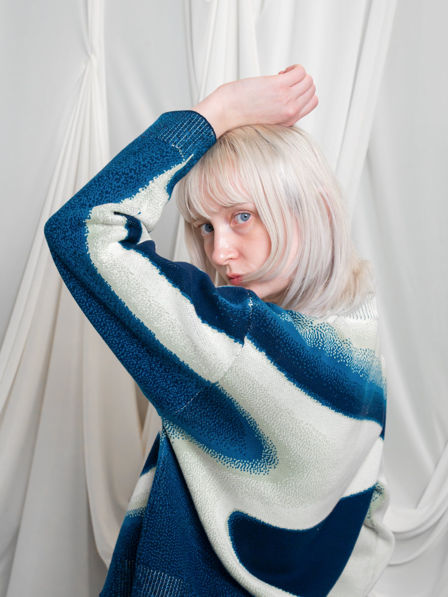 Woman wearing blue and white cropped knit stardust sweater in size extra small posing with hand over head in front of white draped fabric.