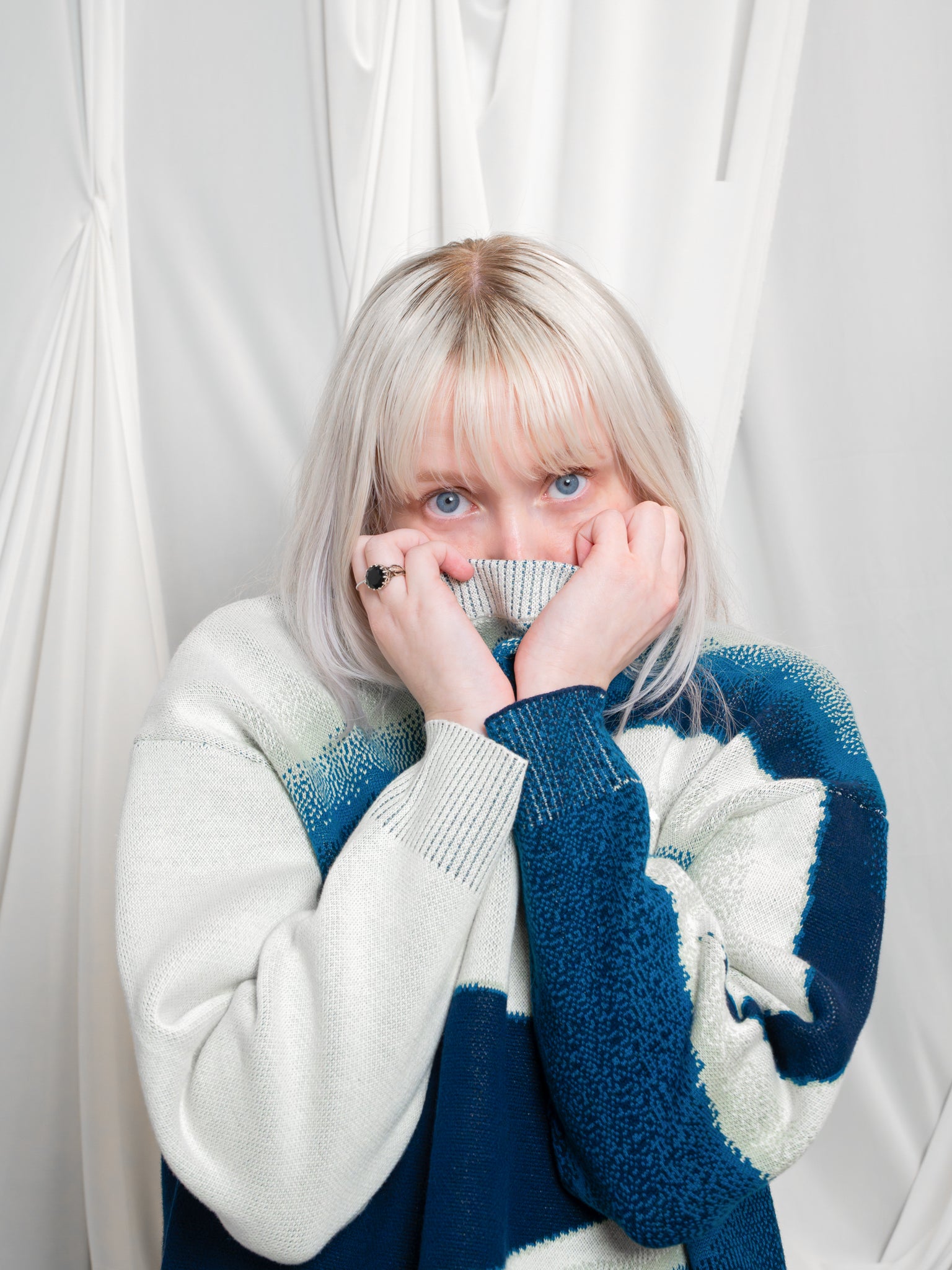 Woman wearing blue and white cropped knit stardust sweater in size extra small holding neckline over her face in front of white draped fabric.