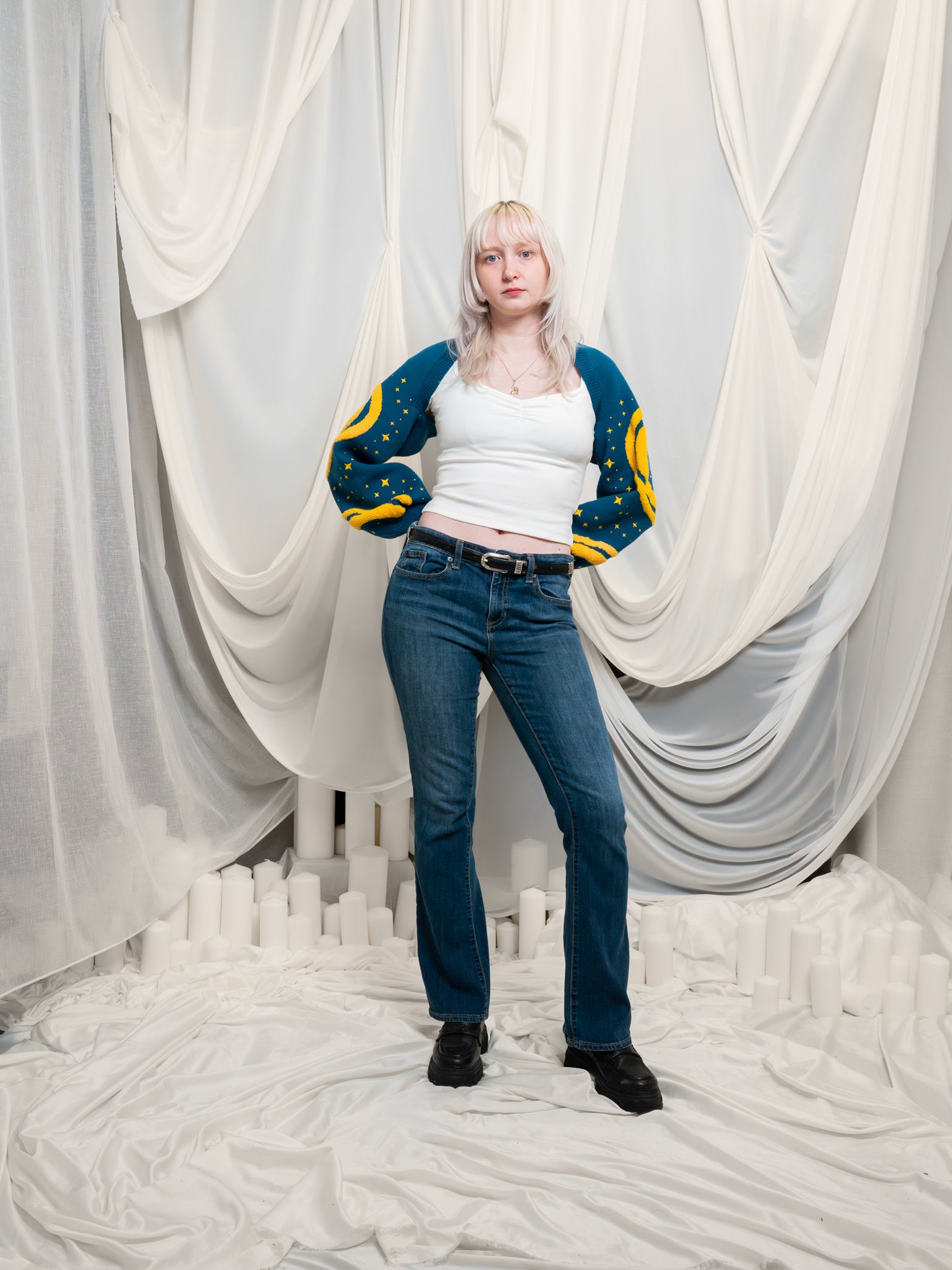 Woman wearing blue and yellow blue moon bolero in size extra small with dark blue jeans and supernova cap sleeve tee in front of draped white fabric.