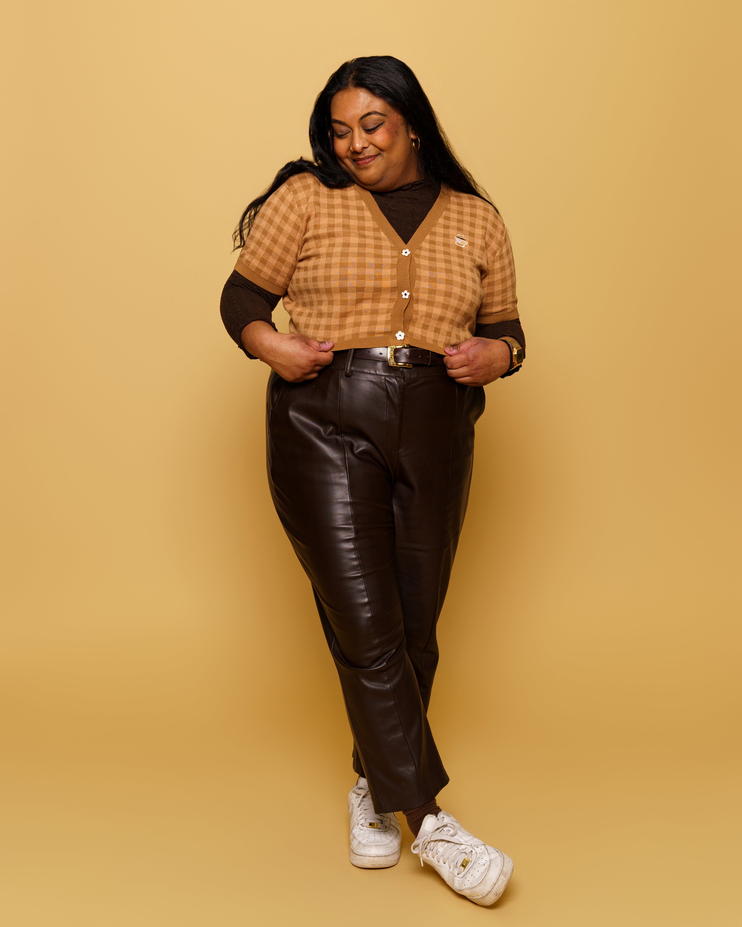Woman wearing brown gingham cappuccino mini cardigan in size 3XL with black pants in front of yellow background.
