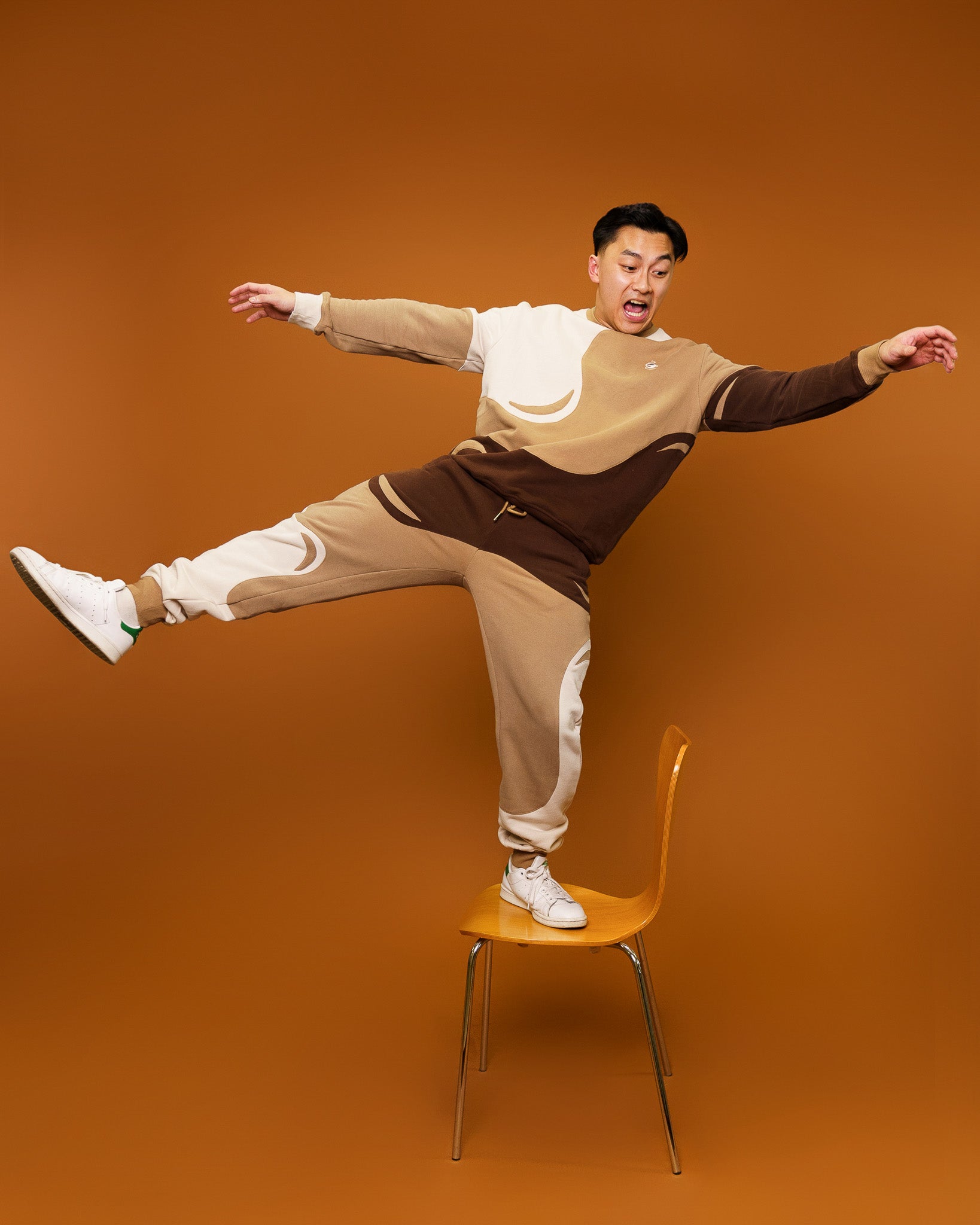 Man wearing brown and white cold-brew crewneck in size small and cold-brew joggers balancing on chair in front of orange background.