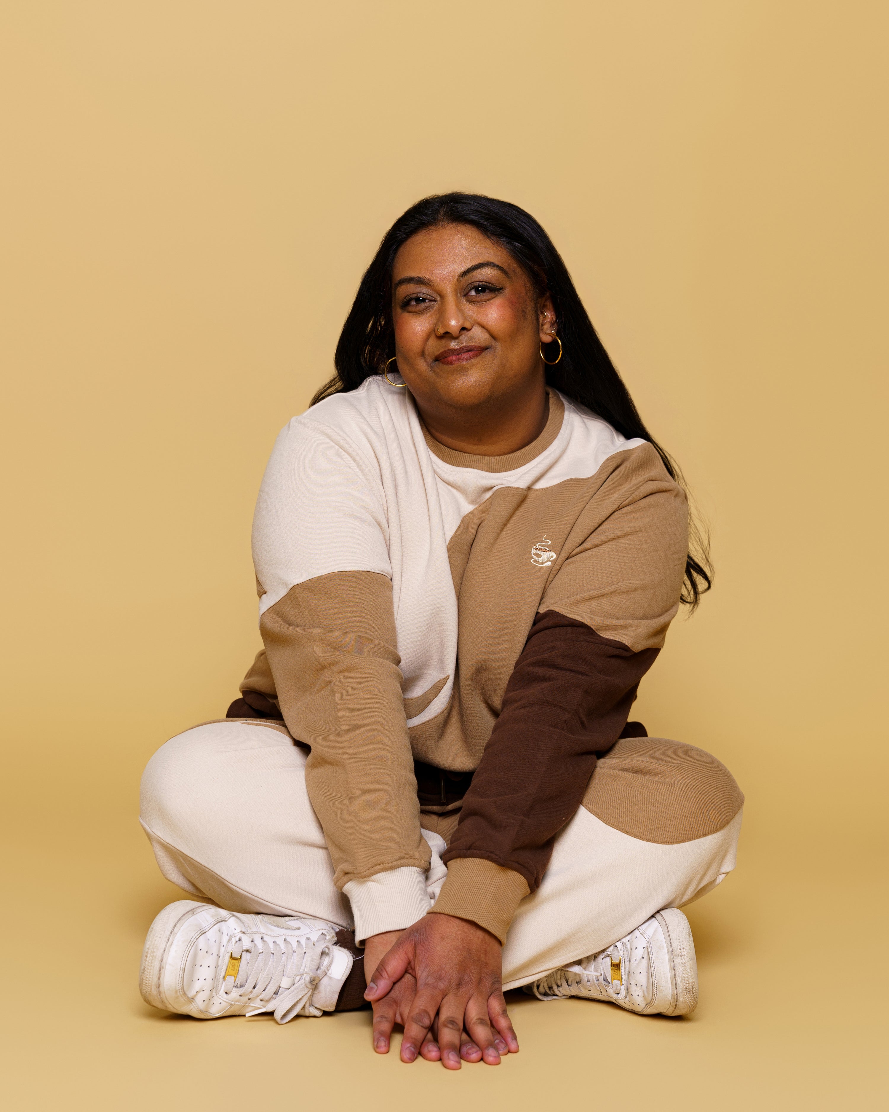 Woman wearing brown and white cold-brew joggers in size 3XL with cold-brew crewneck sitting on floor in front of yellow background.