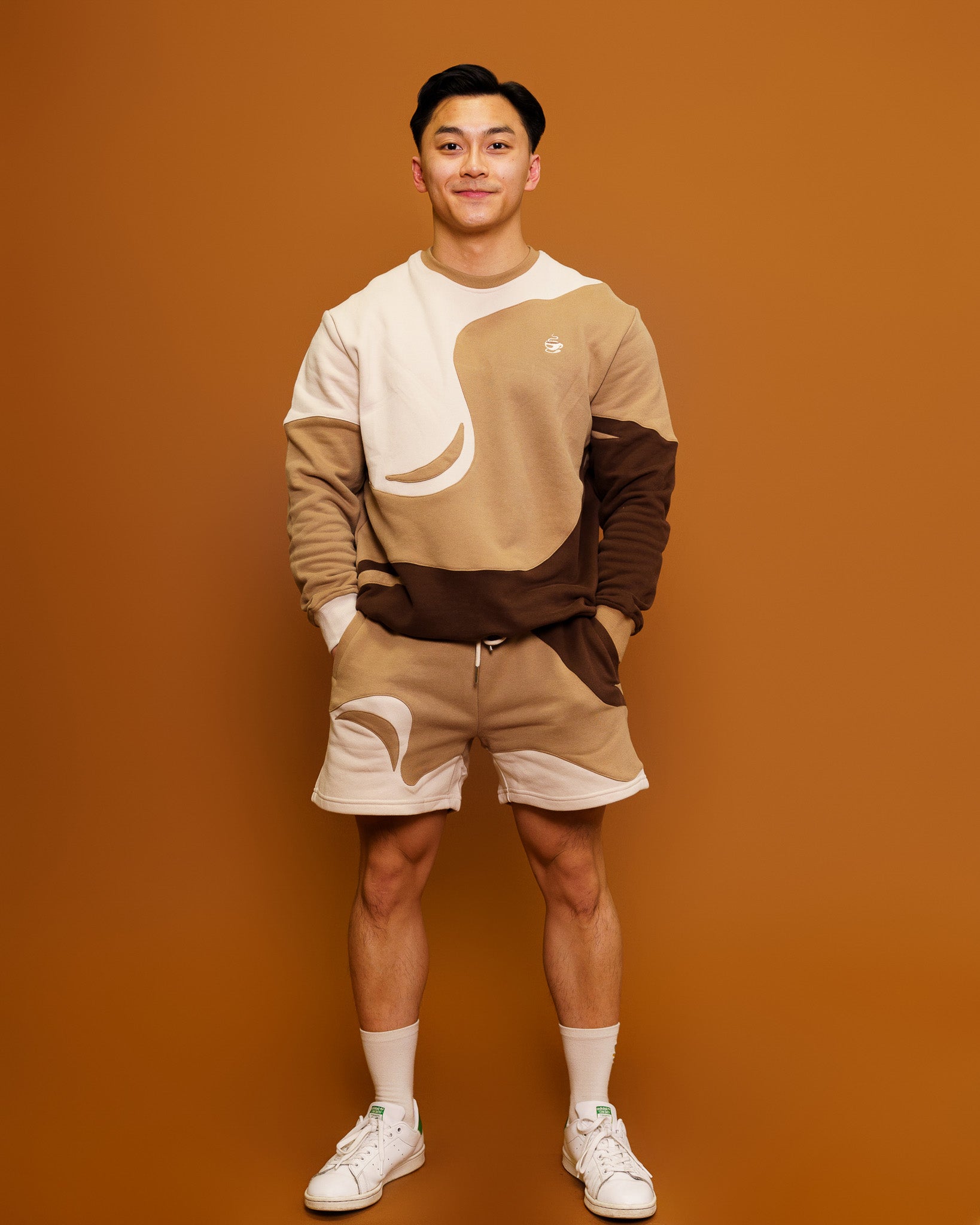 Man wearing brown and white cold-brew shorts in size small and cold-brew crewneck standing in front of orange background.