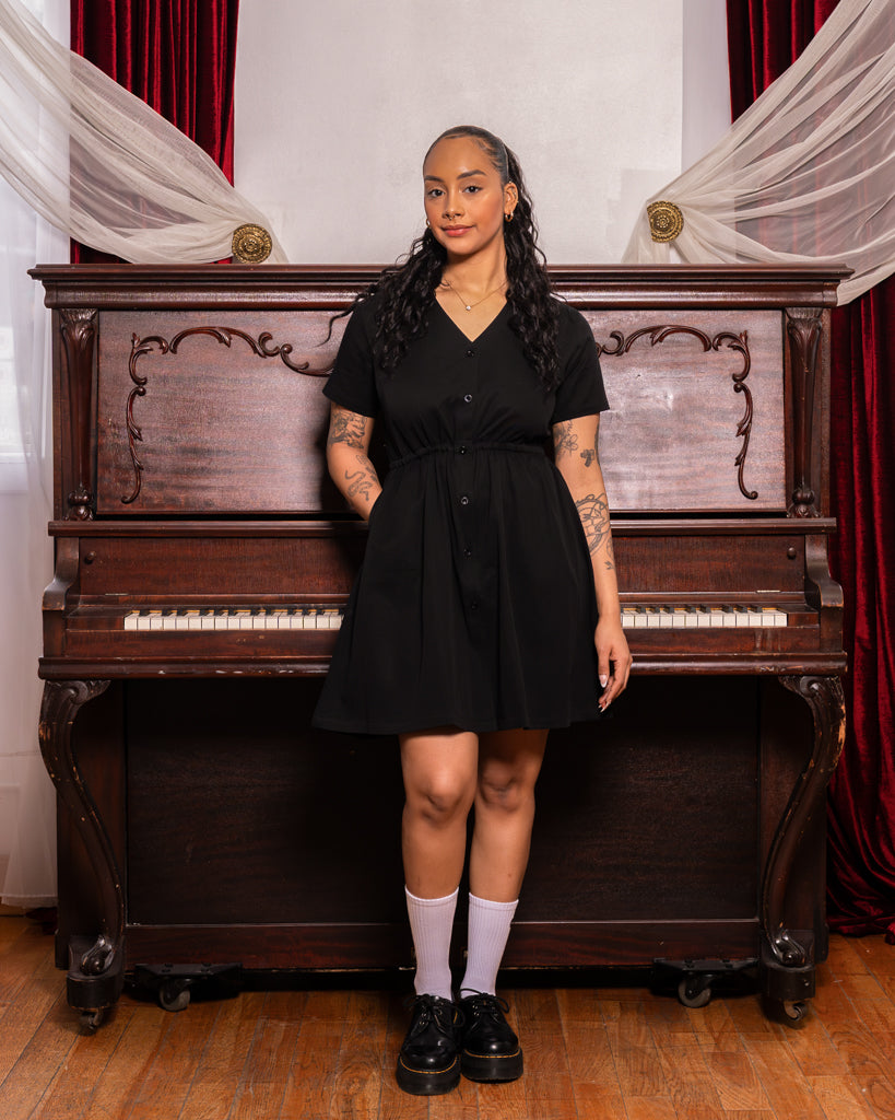 Woman wearing curtsy black smock dress in size small standing in front of piano.