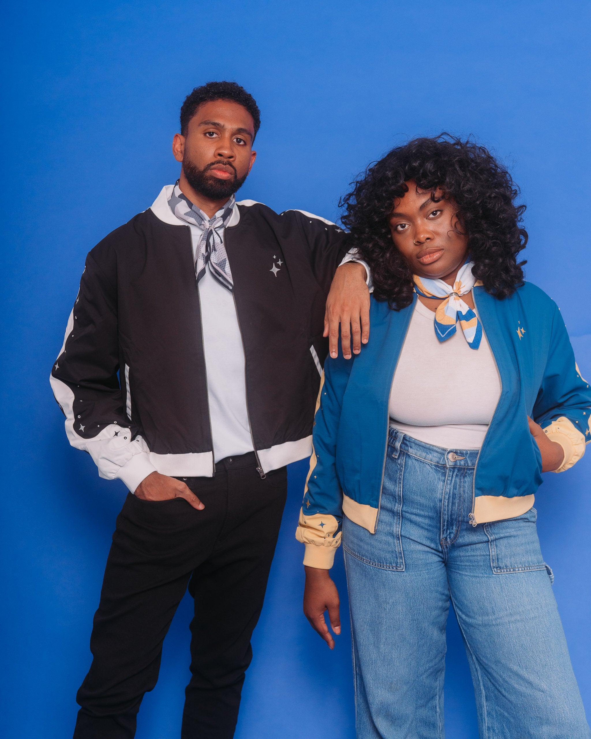 Man wearing deep space bomber jacket in size extra large beside woman wearing blue moon bomber jacket in size medium.
