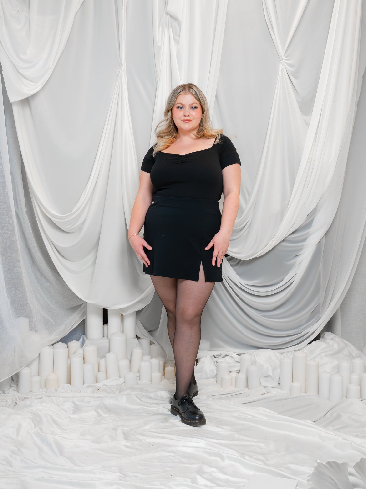 Woman wearing deep space (black) cap sleeve tee in size large with a black skirt and shoes in front of a while, fabric-draped background.