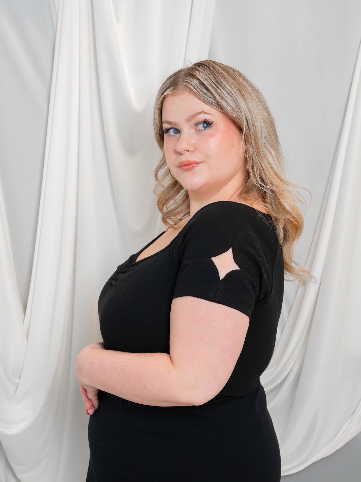 Side profile close-up of woman wearing deep space (black) cap sleeve tee in front of white draped fabric, showcasing the sparkle shaped cut out on the sleeve.