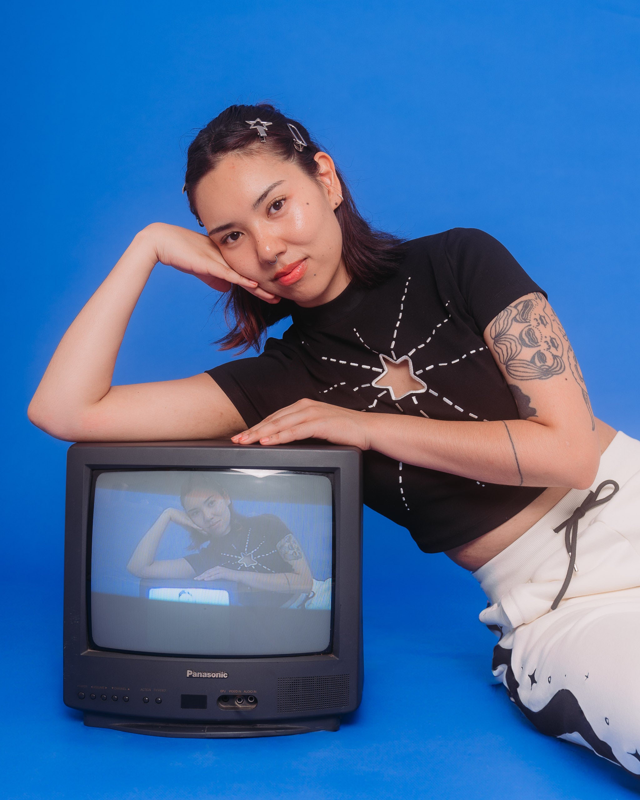 Woman wearing deep space mini tee in size medium leaning on old tv in front of blue background.