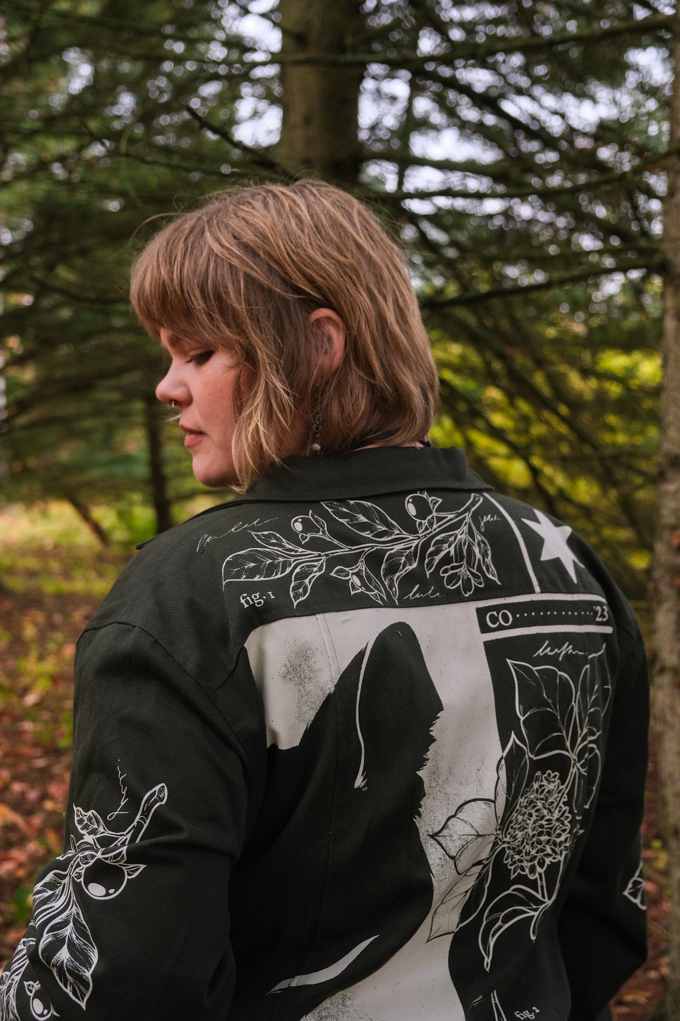 Woman wearing dark green don't cross me jacket in size extra large glancing over the shoulder showing white weathered graphic on back and shoulder.