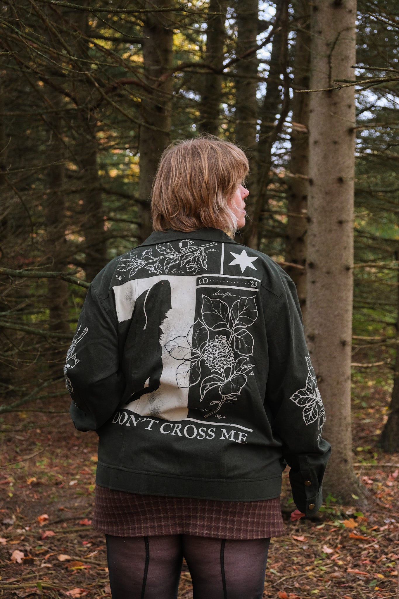 Back view of woman wearing dark green don't cross me jacket in size extra large showcasing white graphic on back featuring flora, half view of a cat, and "Don't Cross Me" text.