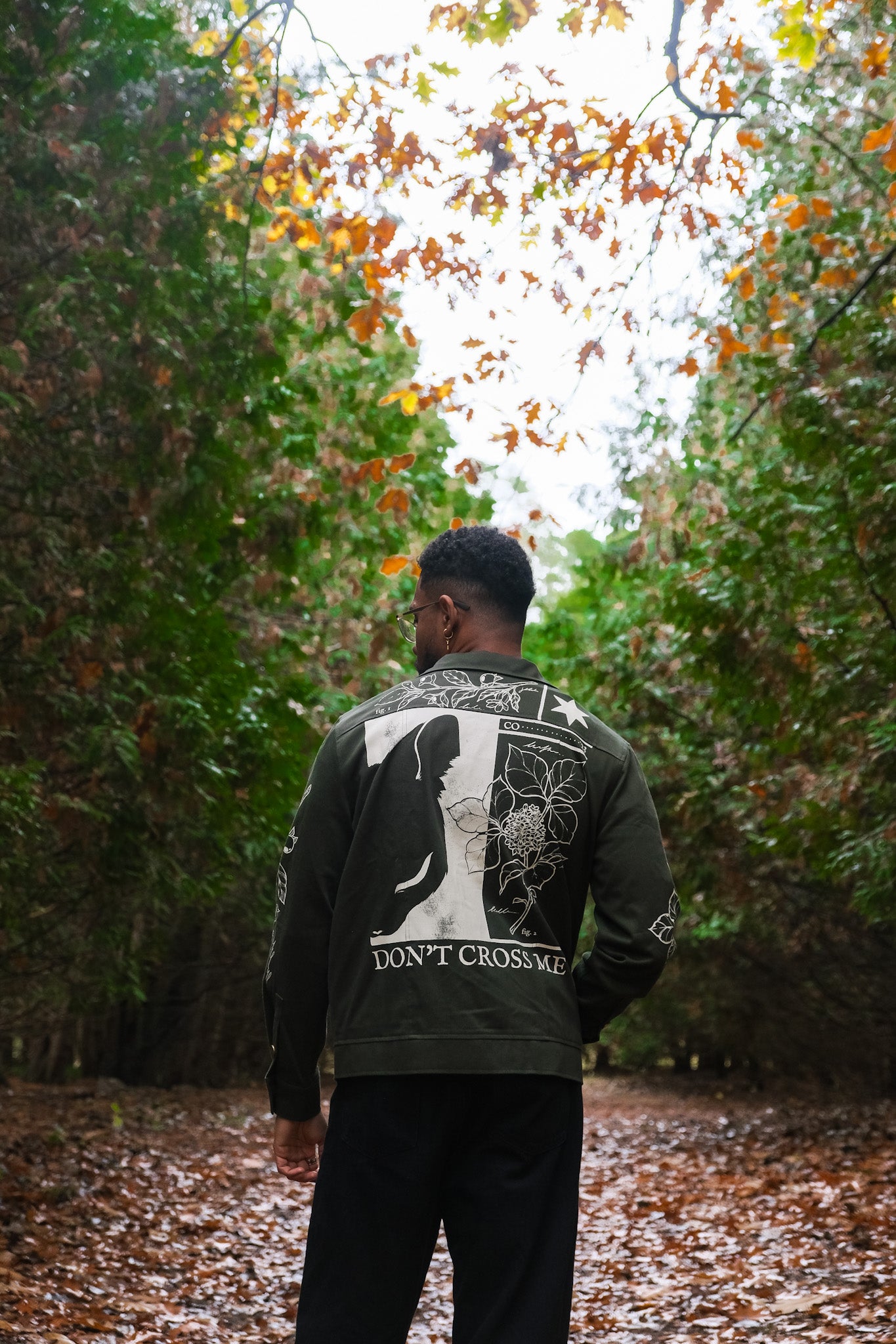 Back view of man wearing dark green don't cross me jacket in size large showing white flora, half of a cat face, and "Don't Cross Me" text.