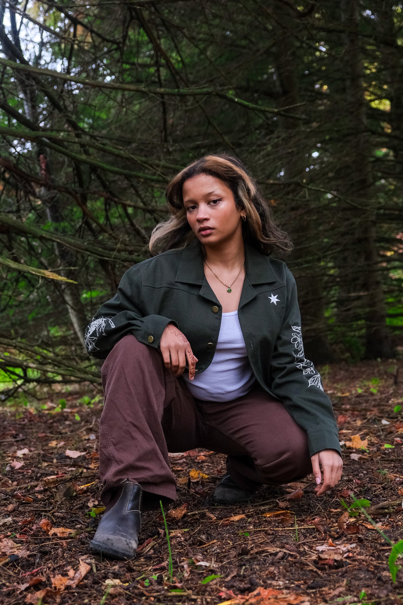 Woman crouched down wearing dark green don't cross me jacket in size extra small in forest.