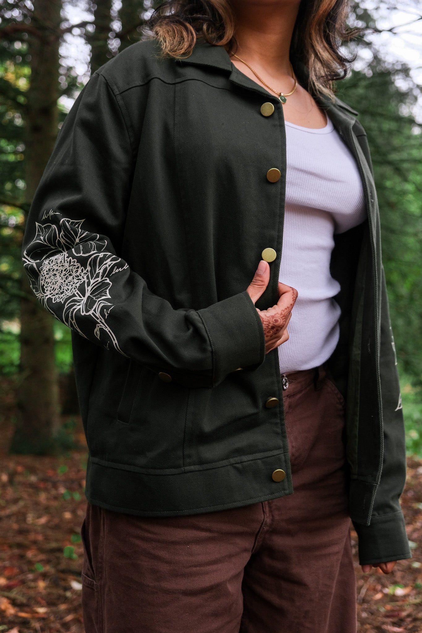 Woman wearing dark green don't cross me jacket in size extra small showing white floral sleeve design in forest.