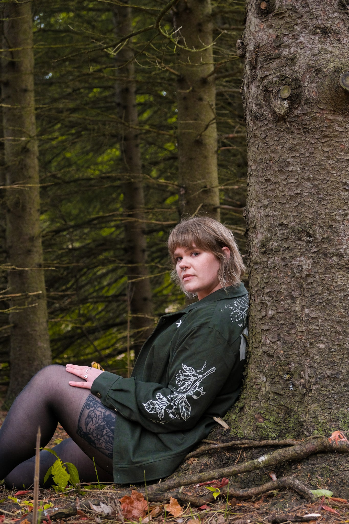 Woman wearing dark green don't cross me jacket in size extra large sitting against tree in forest.