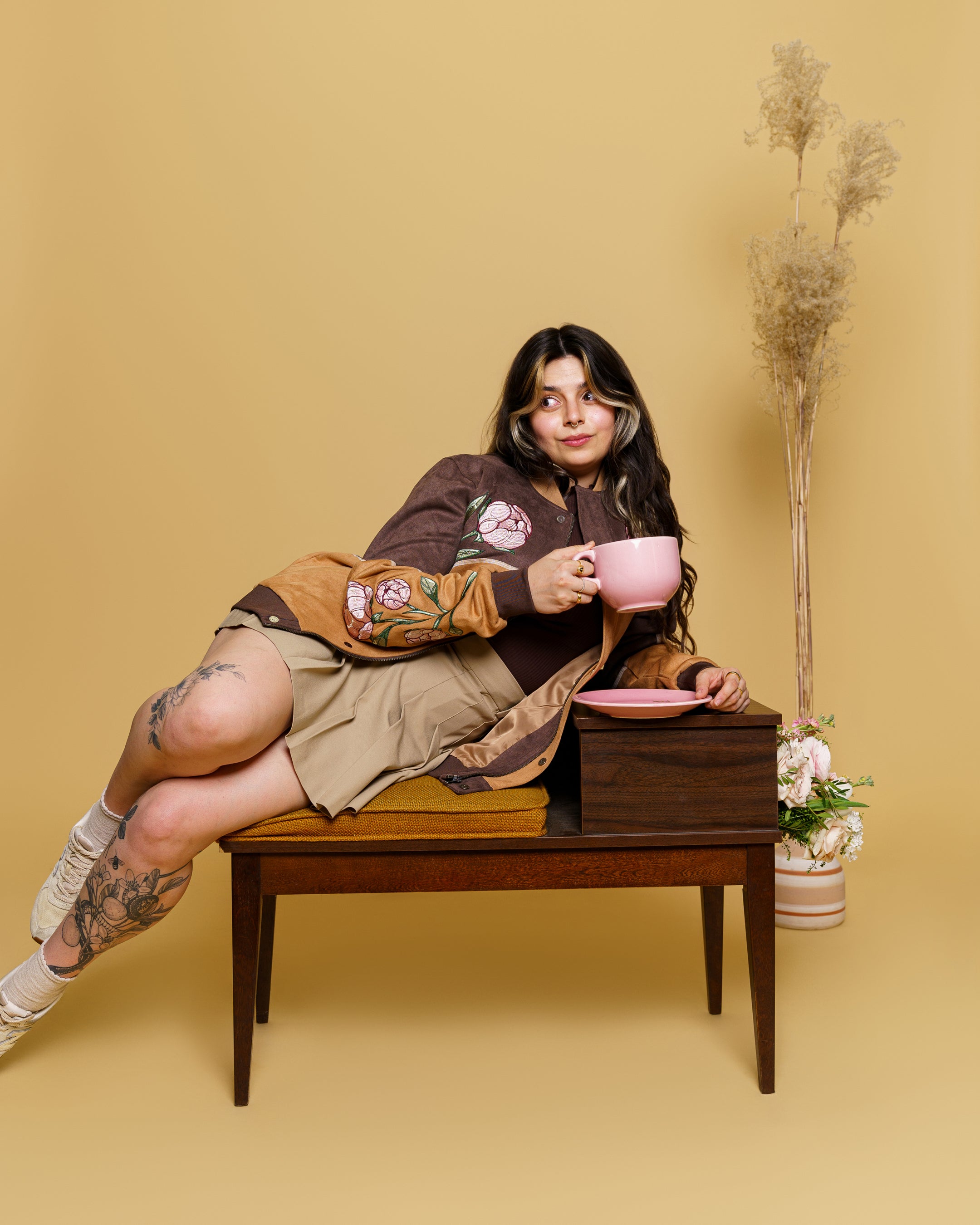 Woman wearing flowers for oscar faux-suede jacket in size small leaning on vintage chair with a pink mug.
