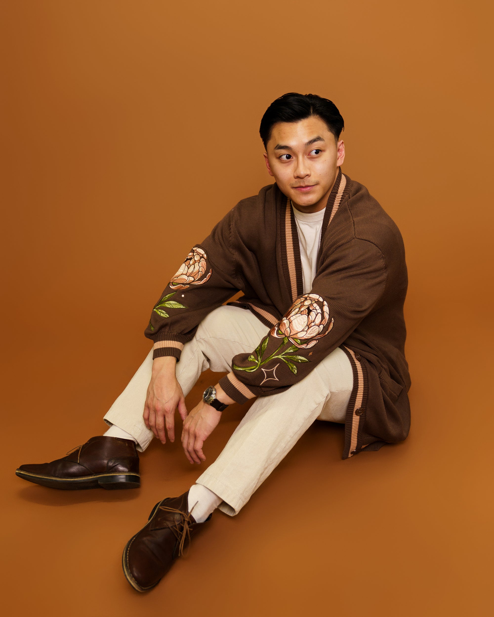 Man wearing freshly picked cardigan in size medium sitting on ground in front of orange background.