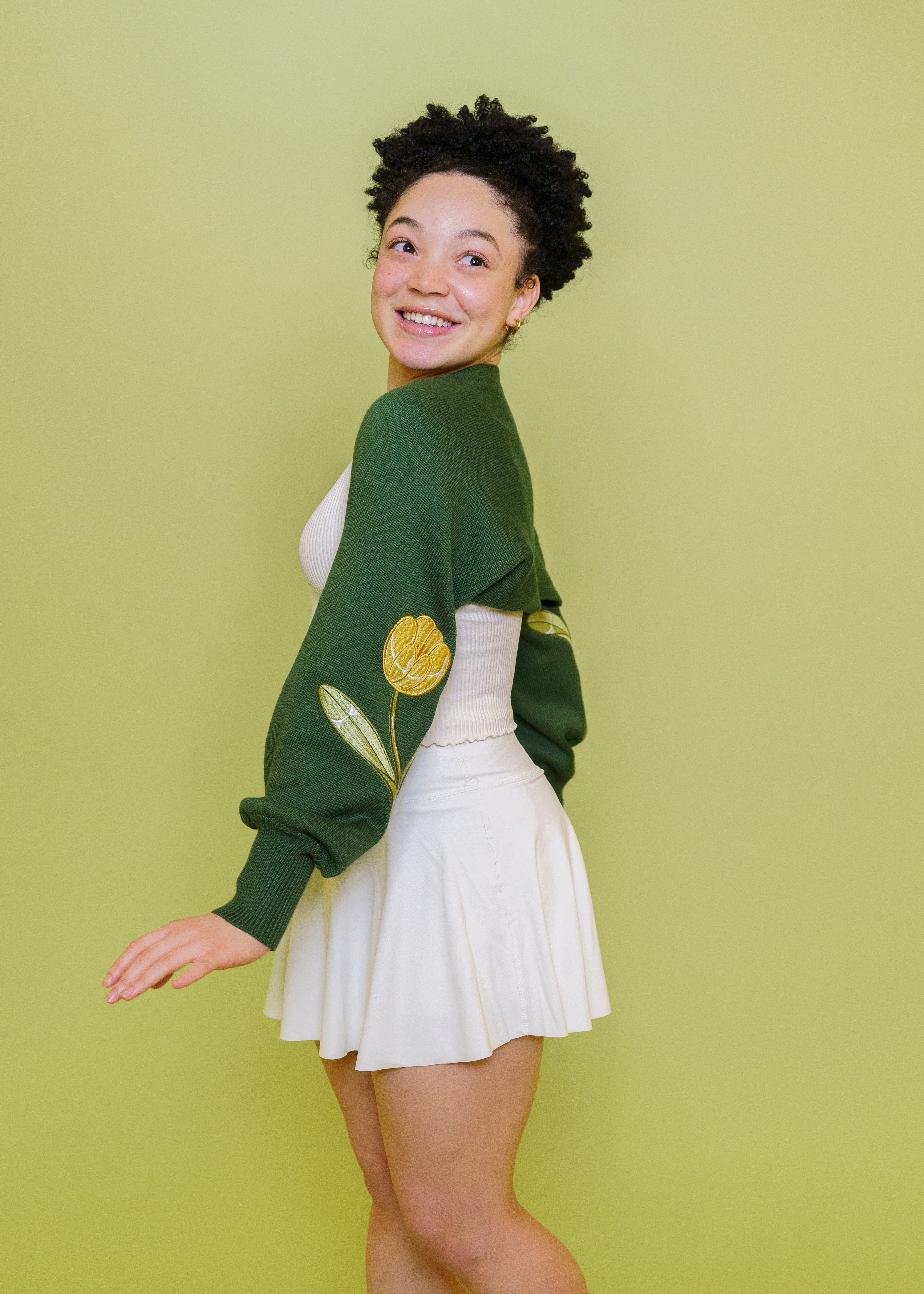 Woman wearing garden green bolero in size extra small glancing over the shoulder in front of green background.