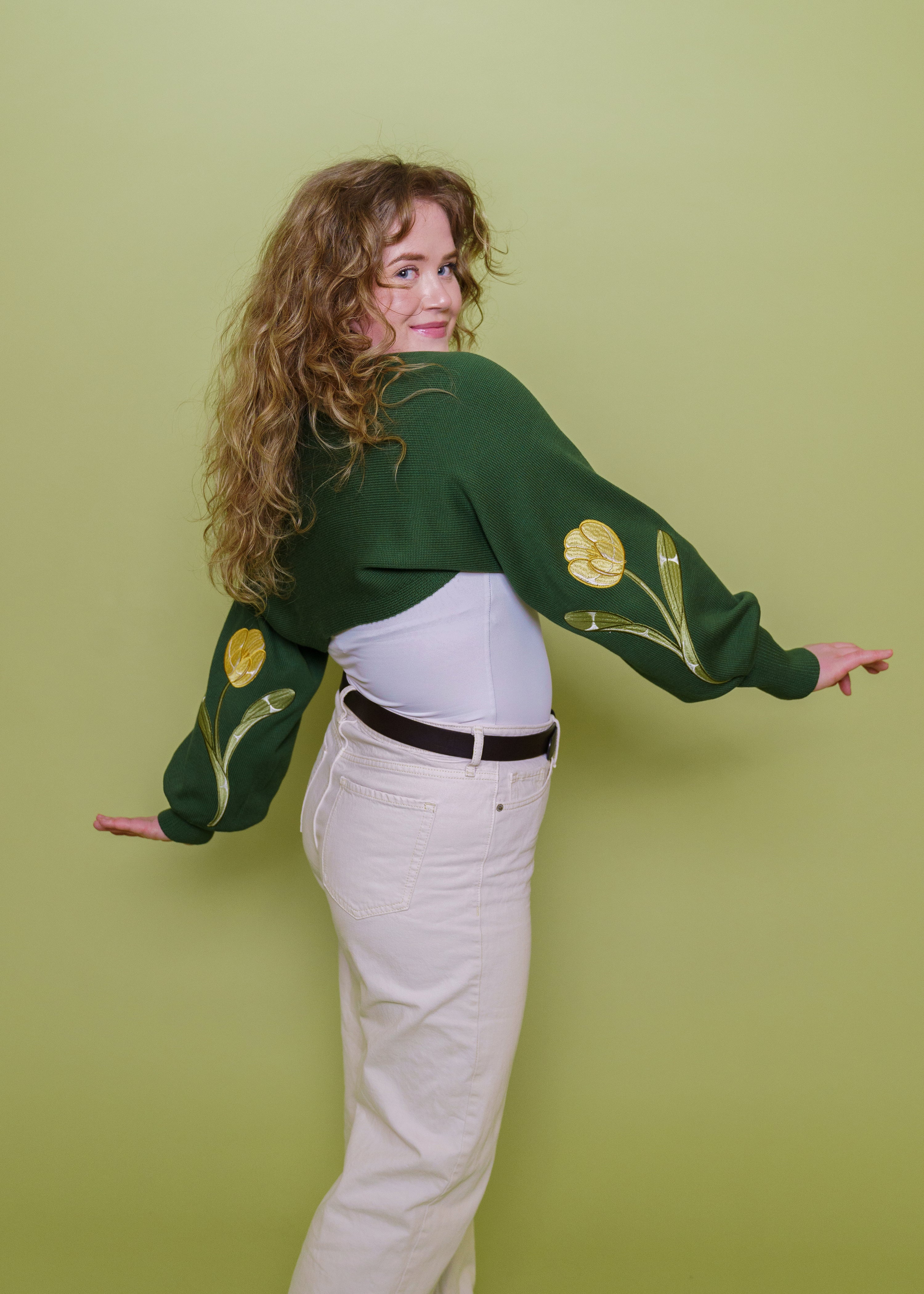 Woman wearing garden green bolero in size medium showing yellow floral embroidery on sleeves in front of green background.