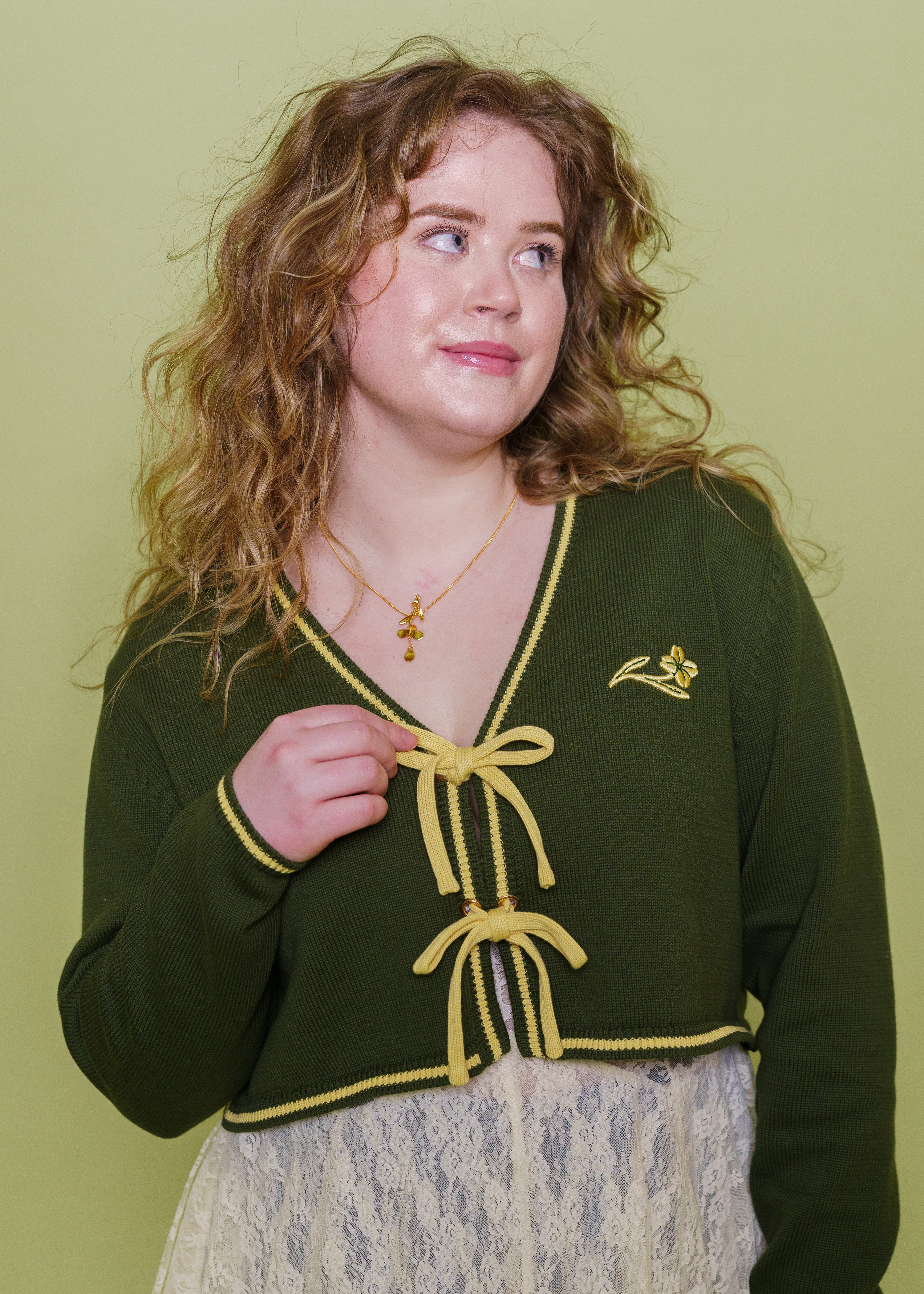 Woman wearing herbal cropped cardigan in size medium holding one of the yellow front closure bows in front of green background.