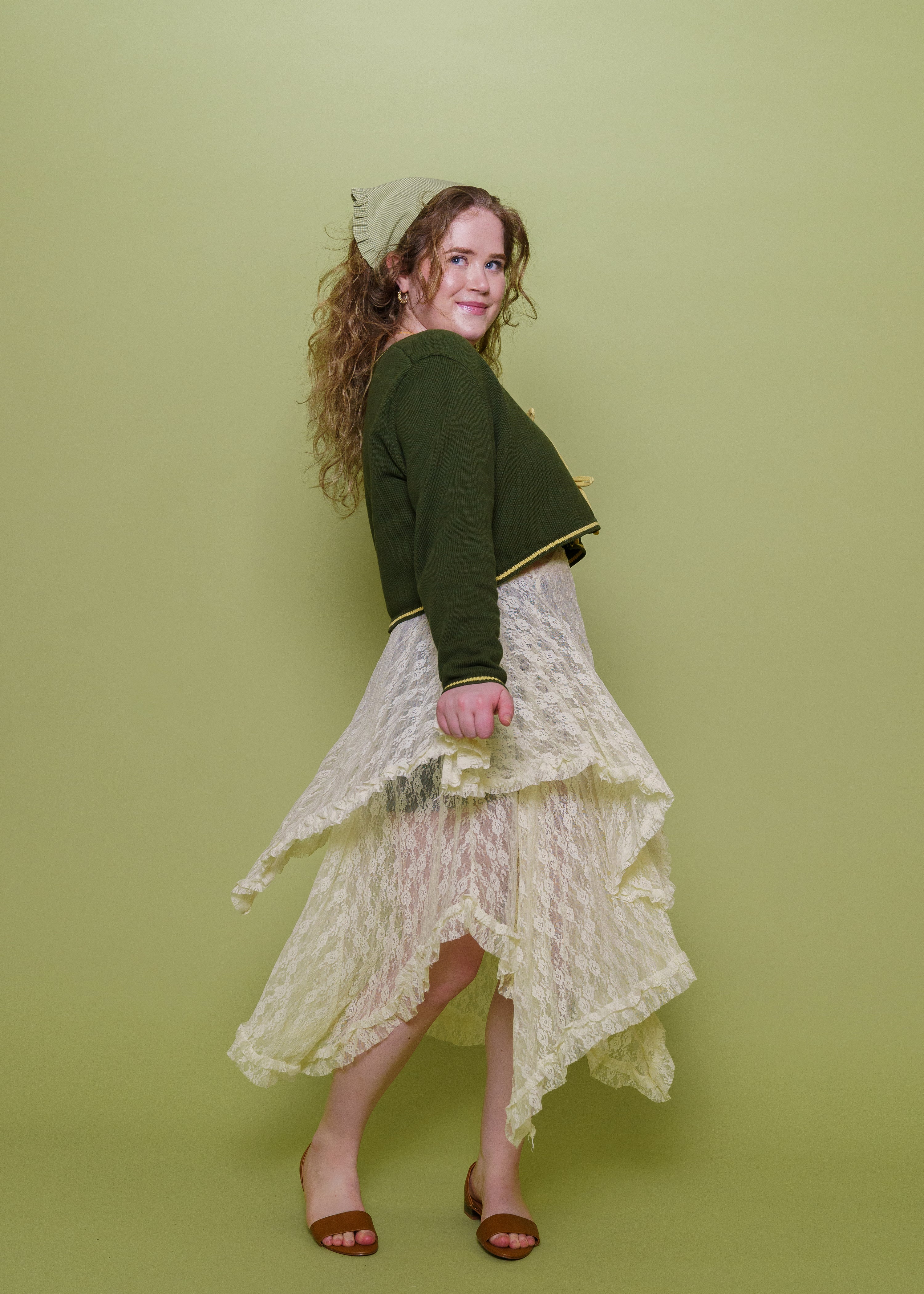 Woman wearing herbal cropped cardigan in size medium with skirt in front of green background.