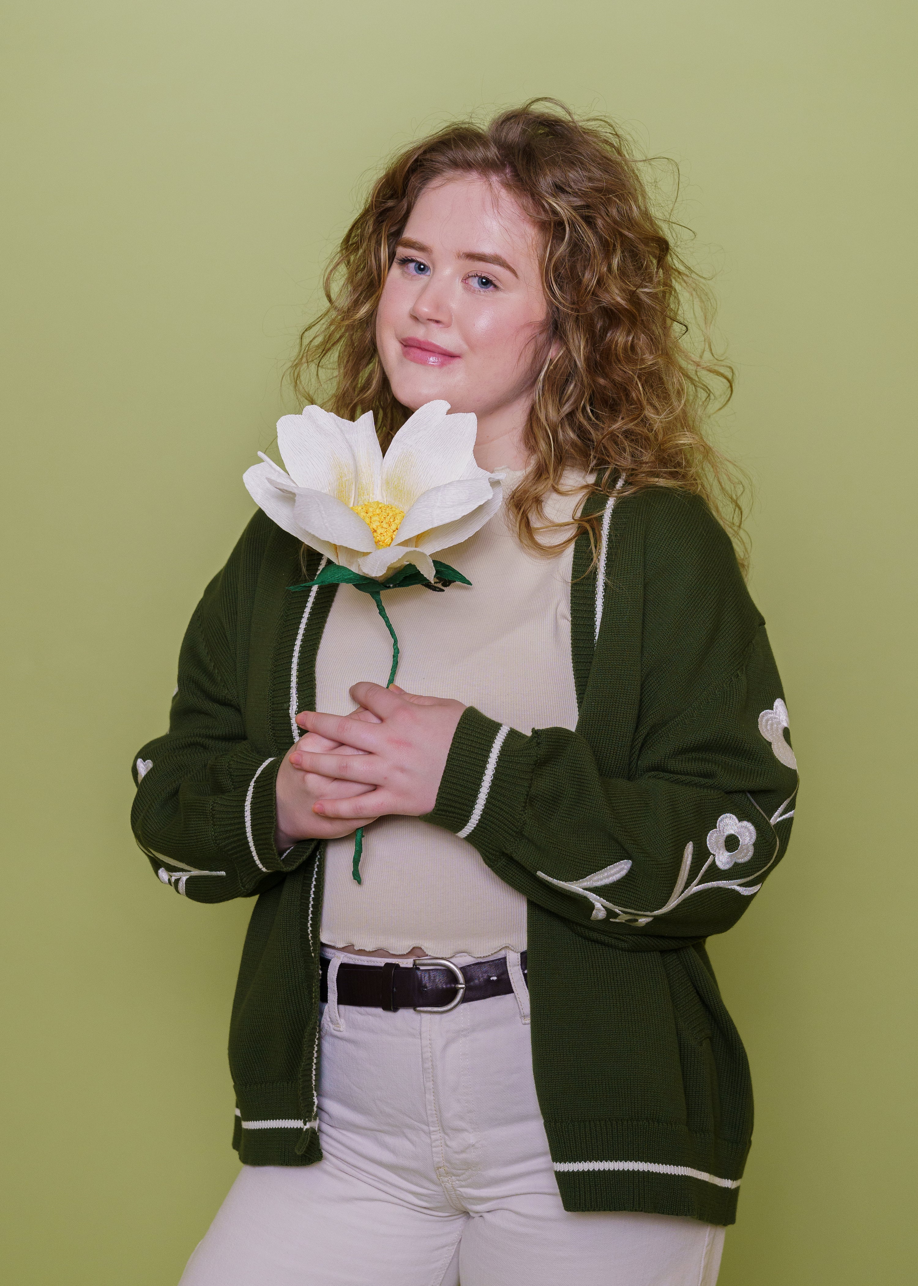 Woman wearing loose leaf cardigan in size large posing with large flower in front of green background.