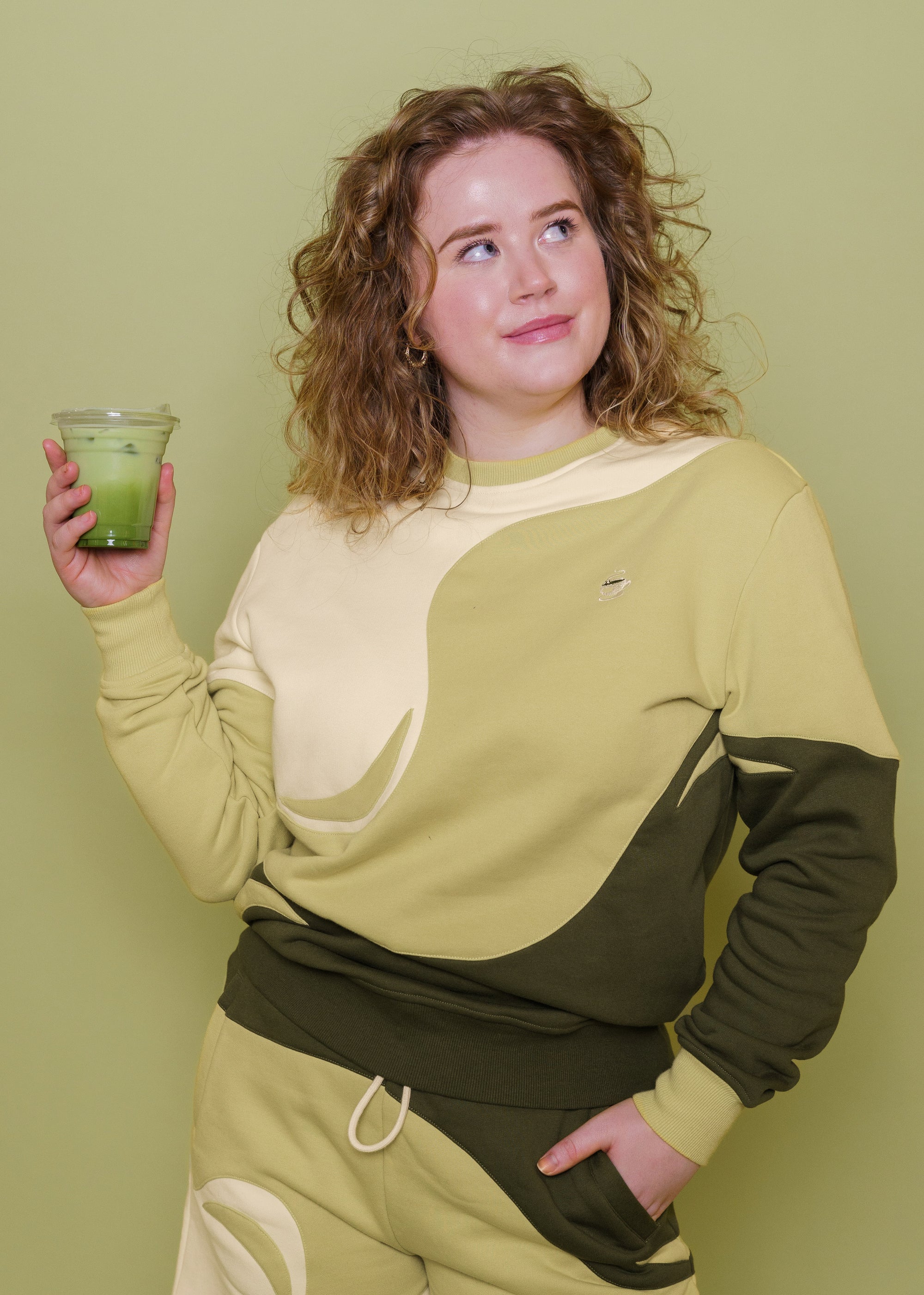 Woman wearing monochromatic green matcha crewneck in size small holding a matcha drink in front of green background.