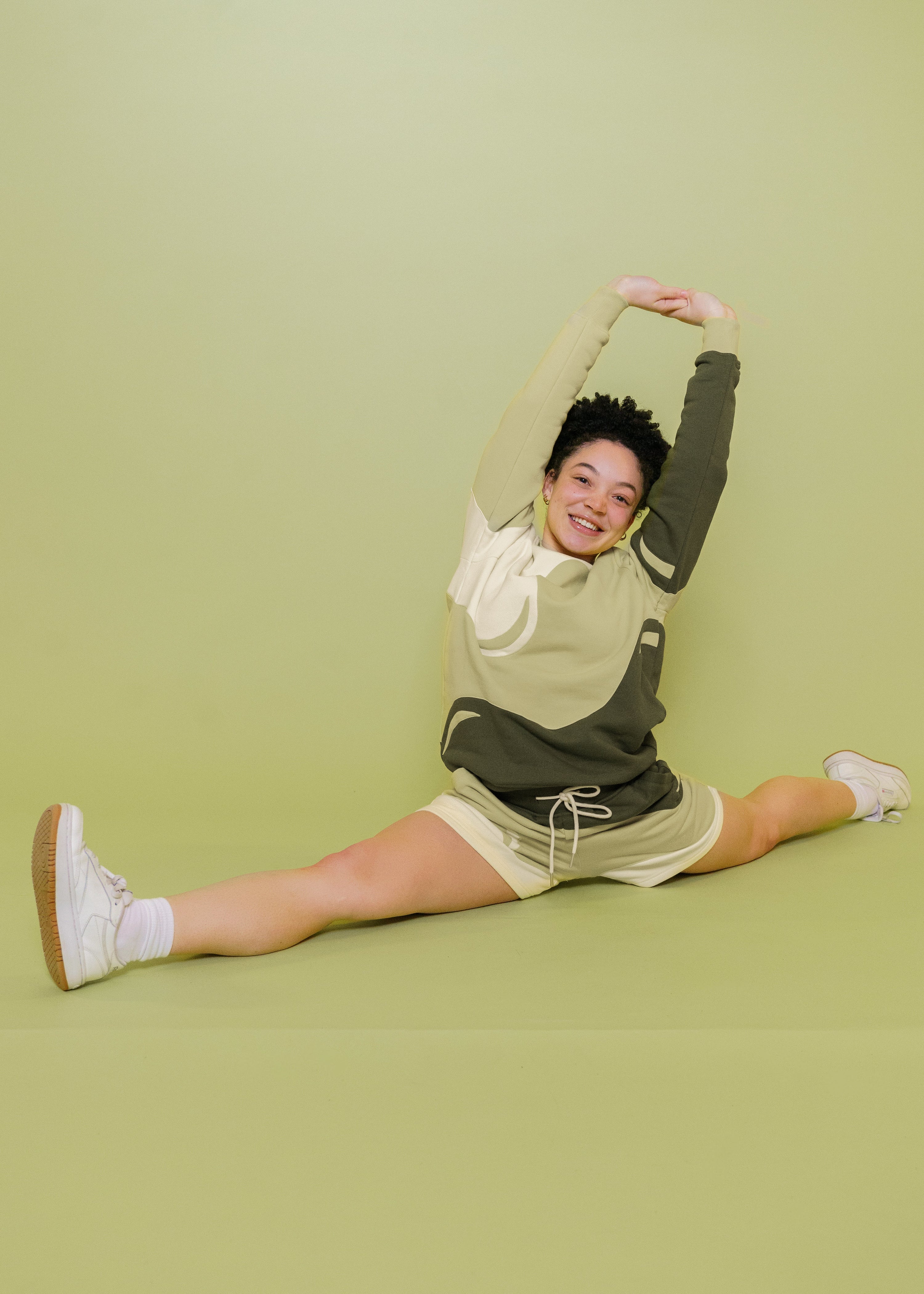 Woman wearing monochromatic green matcha shorts in size extra small with matcha crewneck doing the splits with arms stretched out in front of green background