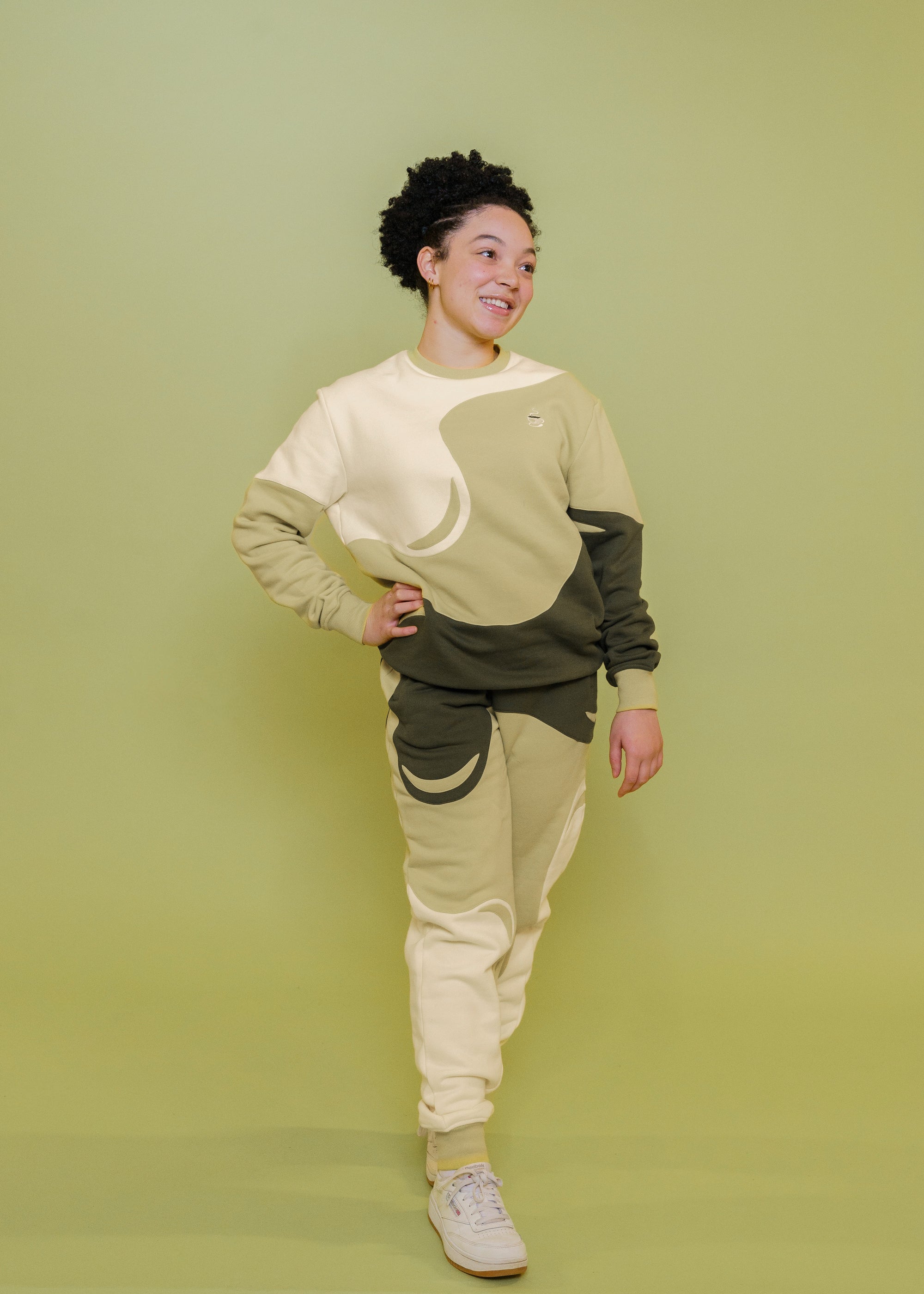 Woman wearing monochromatic green matcha joggers in size extra small and matcha crewneck with white shoes in front of green background.