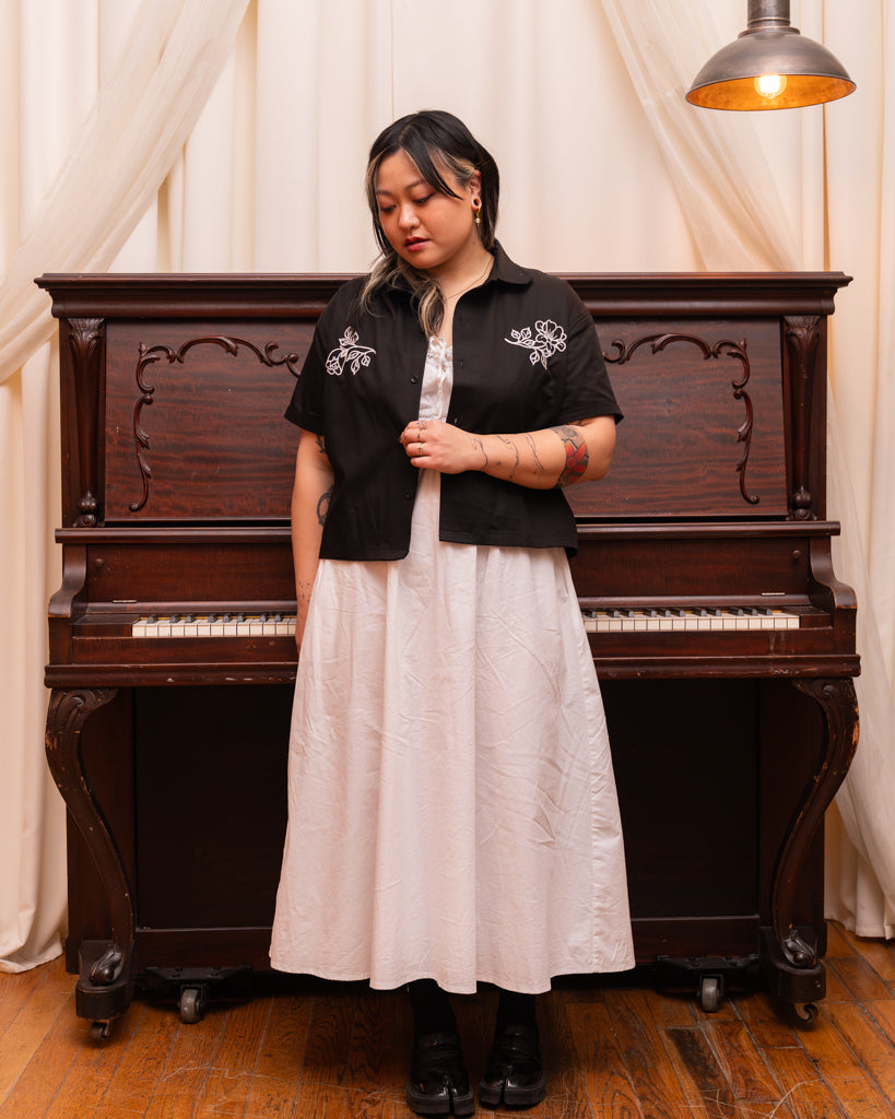 Model wearing mysterious overshirt in size large with white dress standing in front of piano.