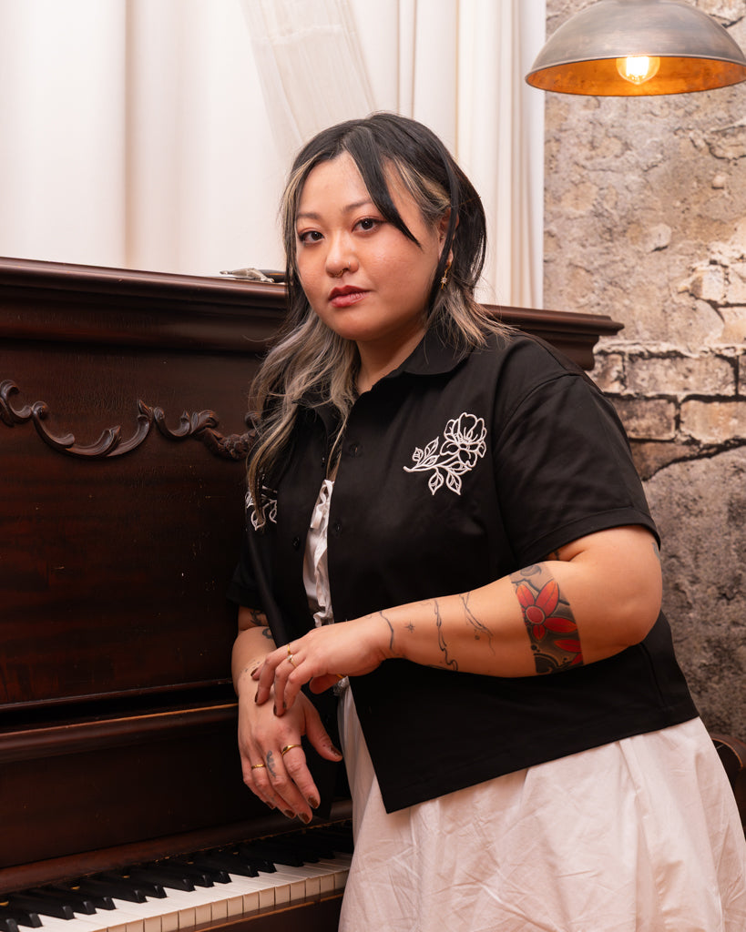 Model wearing mysterious boxy over-shirt in size large with white dress leaning on piano.