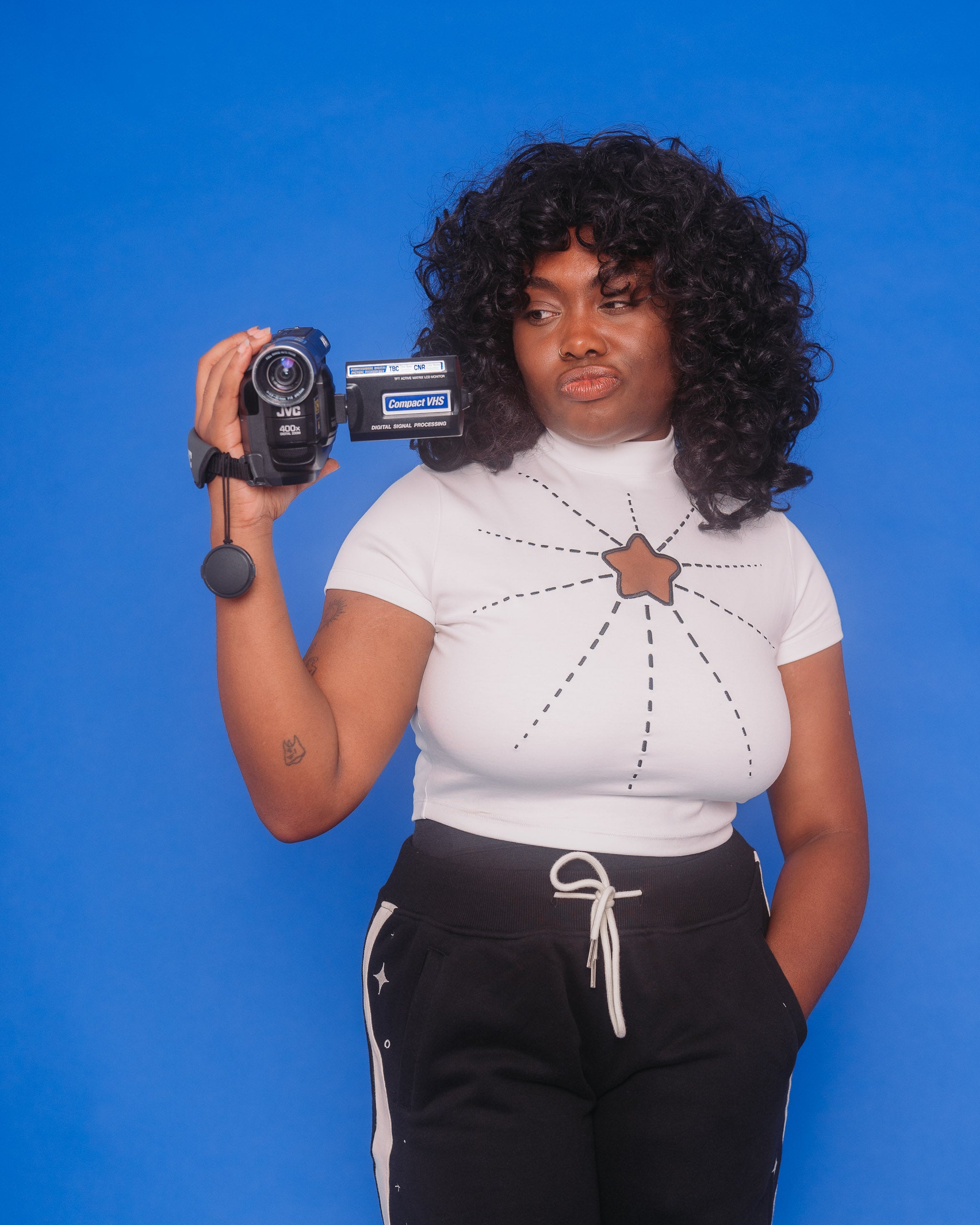 Woman wearing white supernova mini tee in size large holding an old video camera in front of blue background.