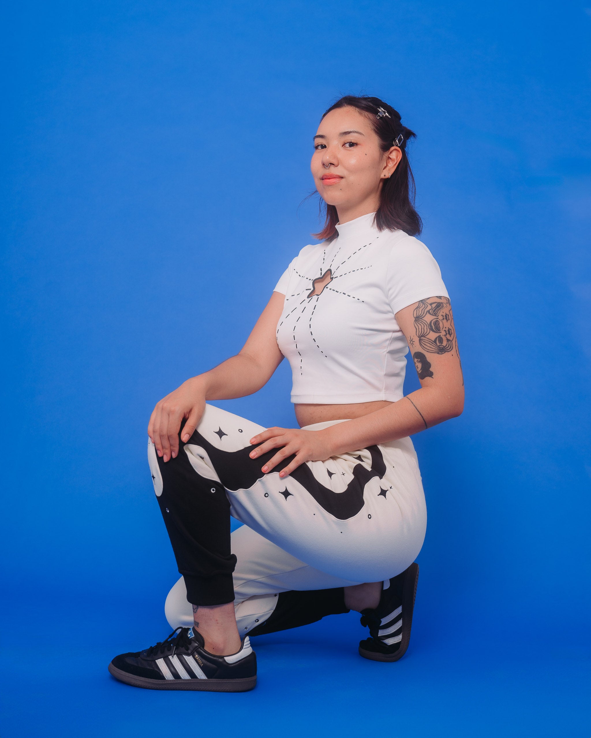 Woman wearing white supernova mini tee in size medium and supernova joggers kneeling down in front of blue background.