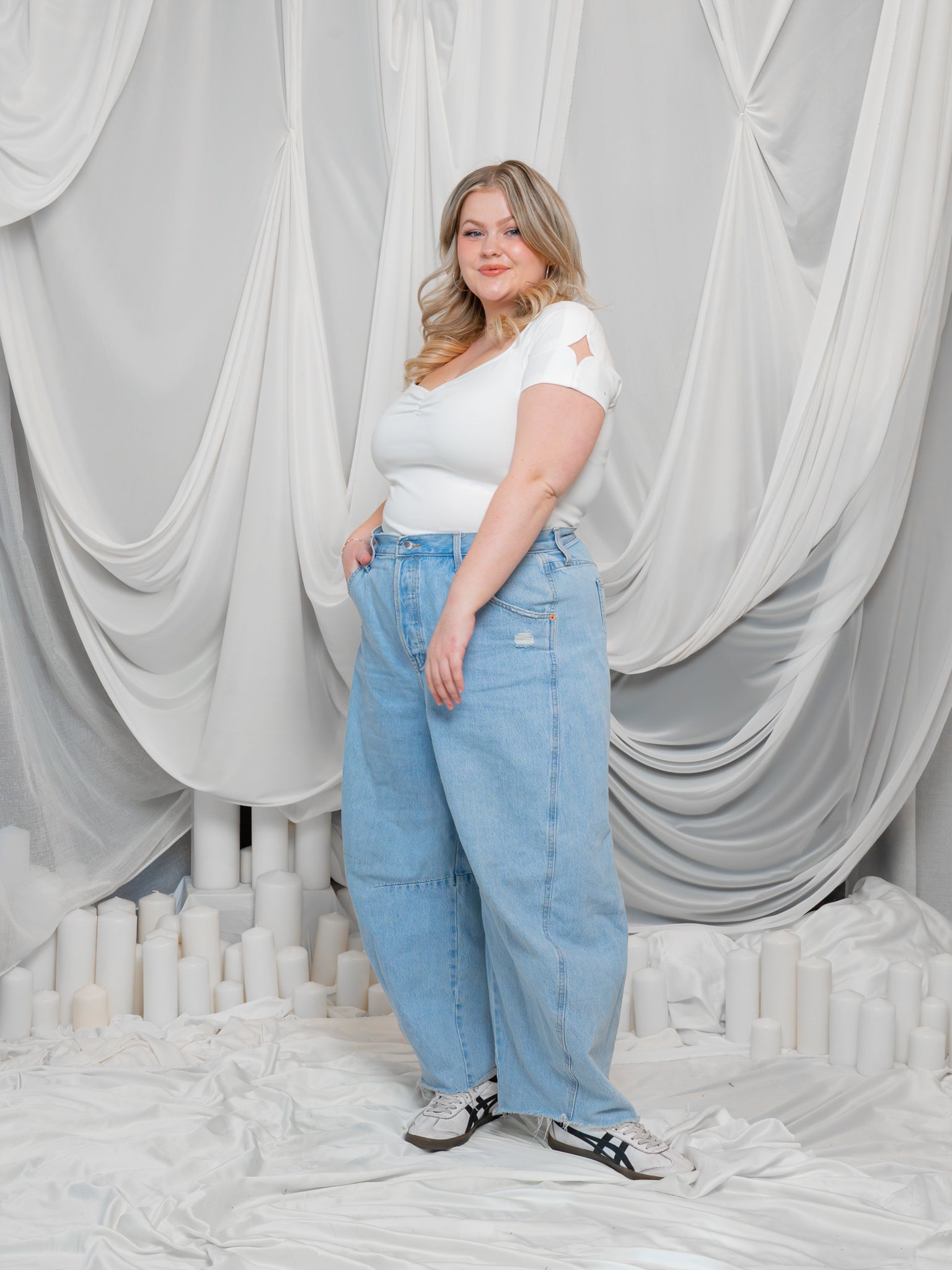 Woman wearing supernova (white) cap sleeve tee with wide leg blue jeans in front of white draped fabric.