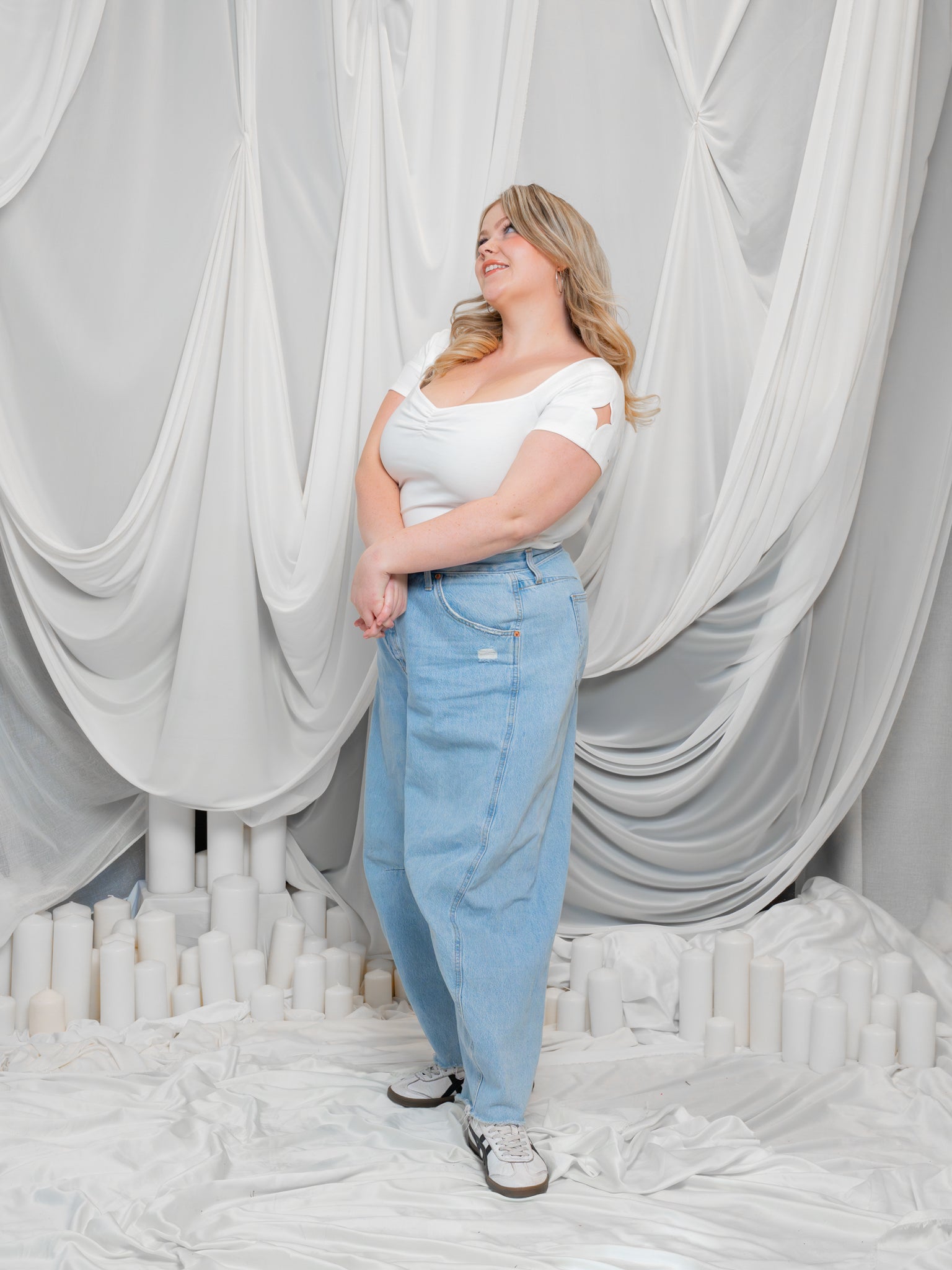 Woman wearing supernova (white) cap sleeve tee in size large with wide leg blue jeans posing in front of white draped fabric.
