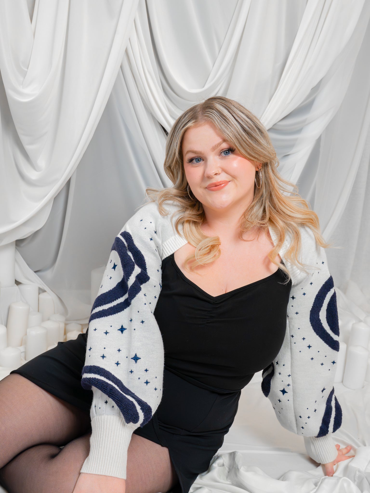 Woman posing wearing dark blue and white supernova bolero in size large in front of white draped fabric.